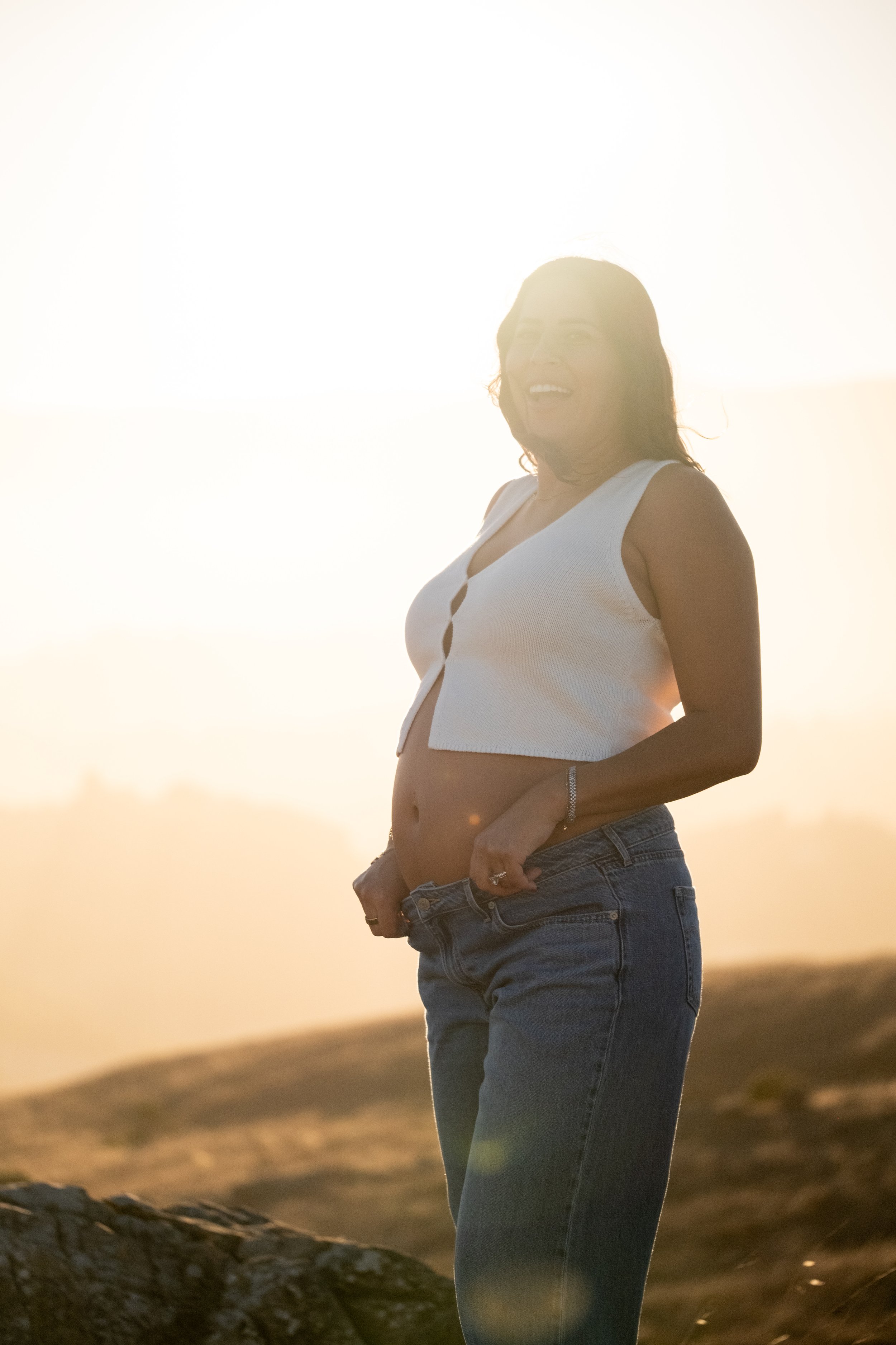 A pregnant woman standing outdoors during sunset, smiling, wearing a white sleeveless top and jeans, with a landscape in the background from a maternity photoshoot in Marin County.