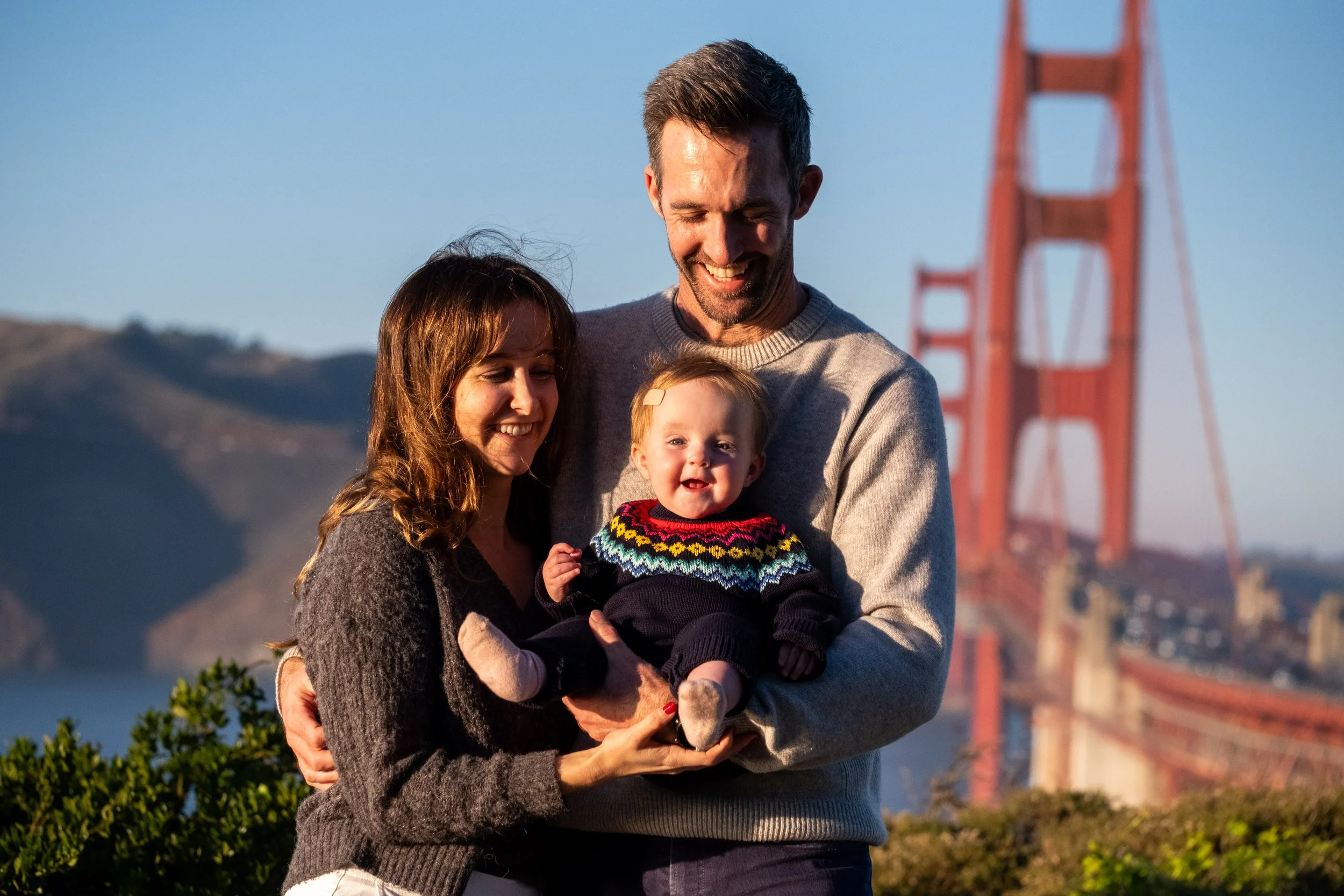 A family of three, a man, a woman, and a young child, smiling and holding each other outdoors with the Golden Gate Bridge in the background during a family photoshoot.