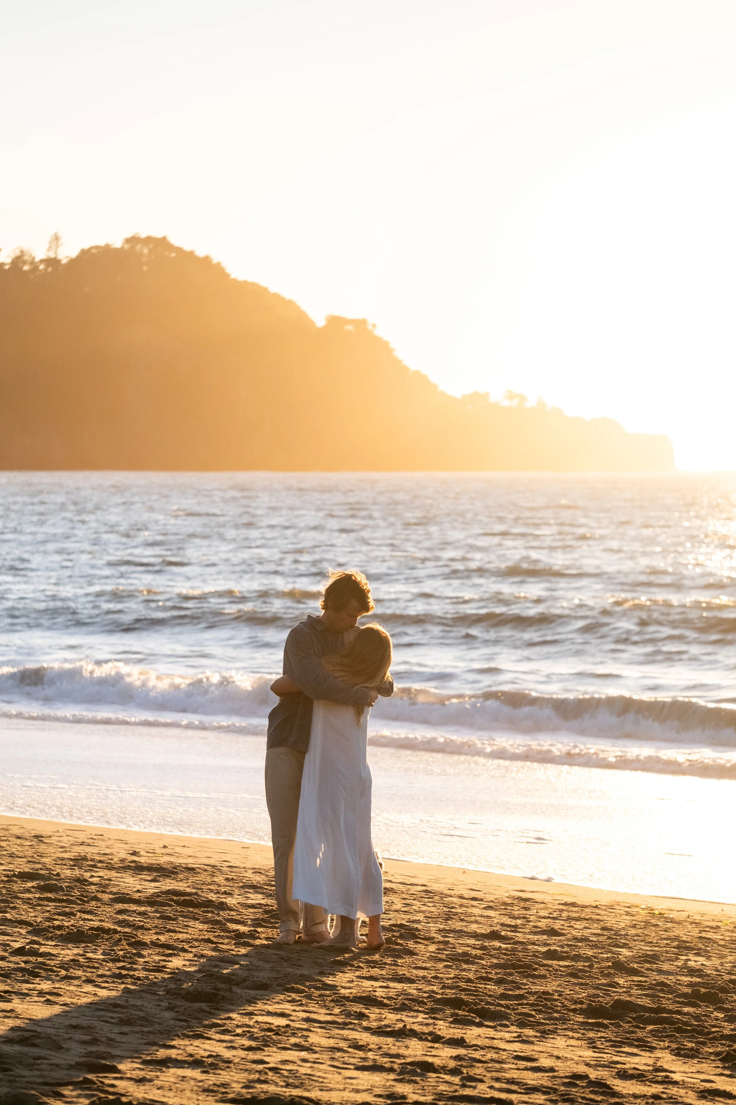 A couple stands on a sandy beach at sunset, embracing each other with the ocean waves behind them and a scenic hillside in the distance, during an engagement photoshoot at Baker Beach in San Francisco.