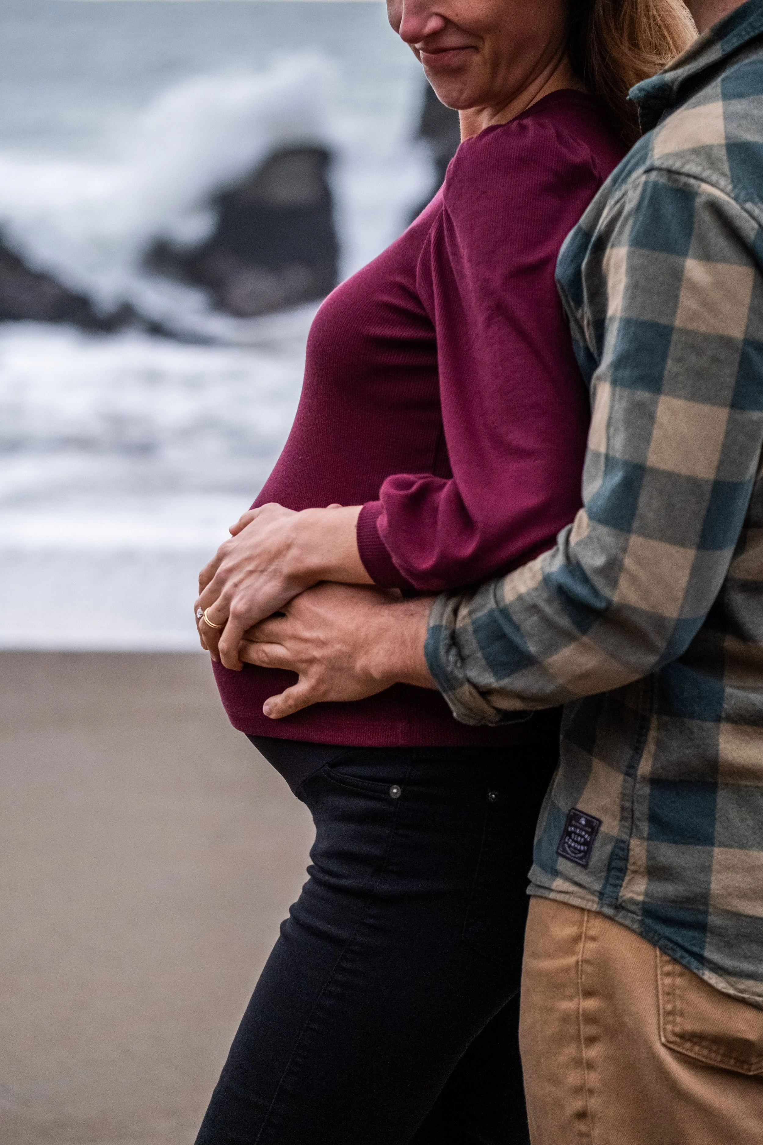 A pregnant woman with reddish hair wearing a burgundy long-sleeve shirt and black pants, standing close to her husband with a plaid shirt and brown pants on a beach, with waves and rocks in the background during a maternity photoshoot.