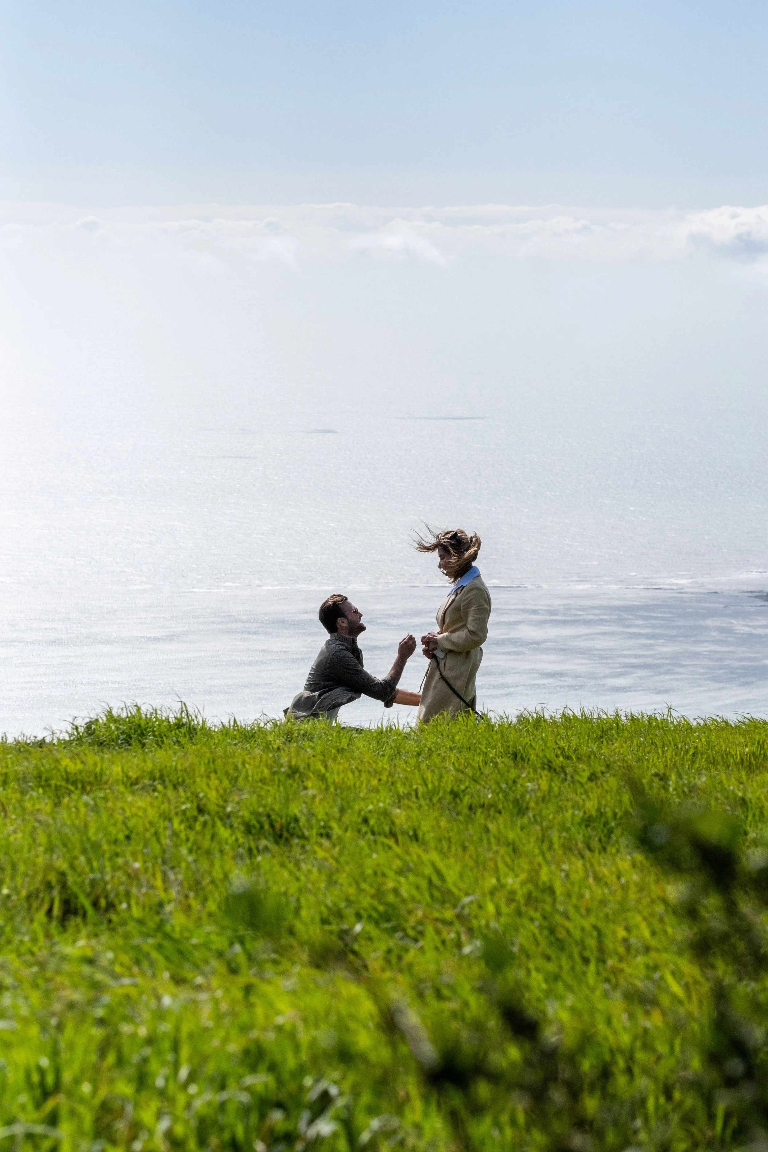 A couple on a grassy field near a body of water, with one person kneeling and proposing marriage to the other, during a proposal photoshoot.