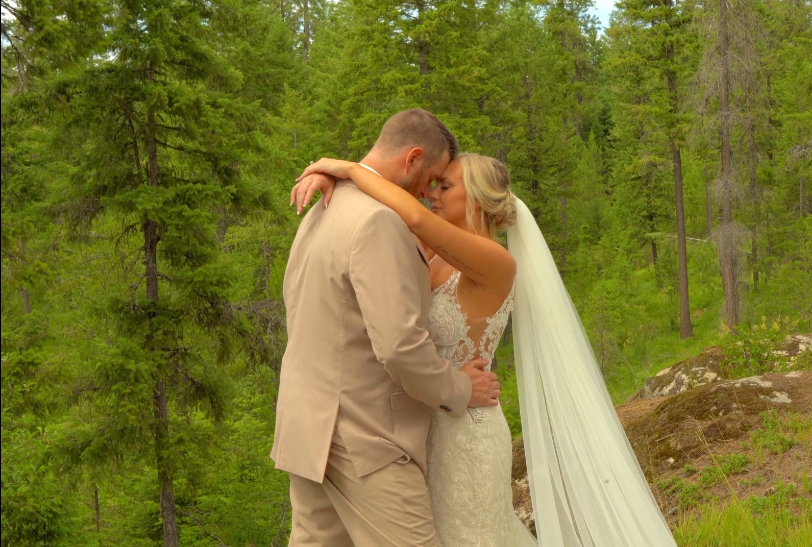 A bride and groom embrace outdoors in a forested area during their wedding, with trees and greenery in the background.