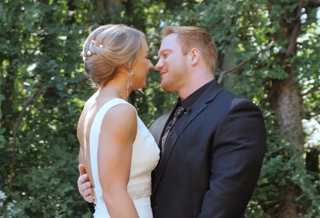 A bride and groom stand close together outdoors, smiling with foreheads touching, surrounded by green trees.