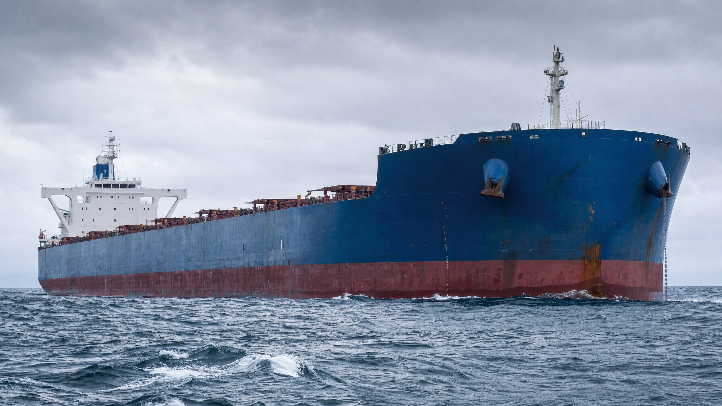 A large cargo ship with a red bottom and blue upper structure sailing on the ocean under a cloudy sky.