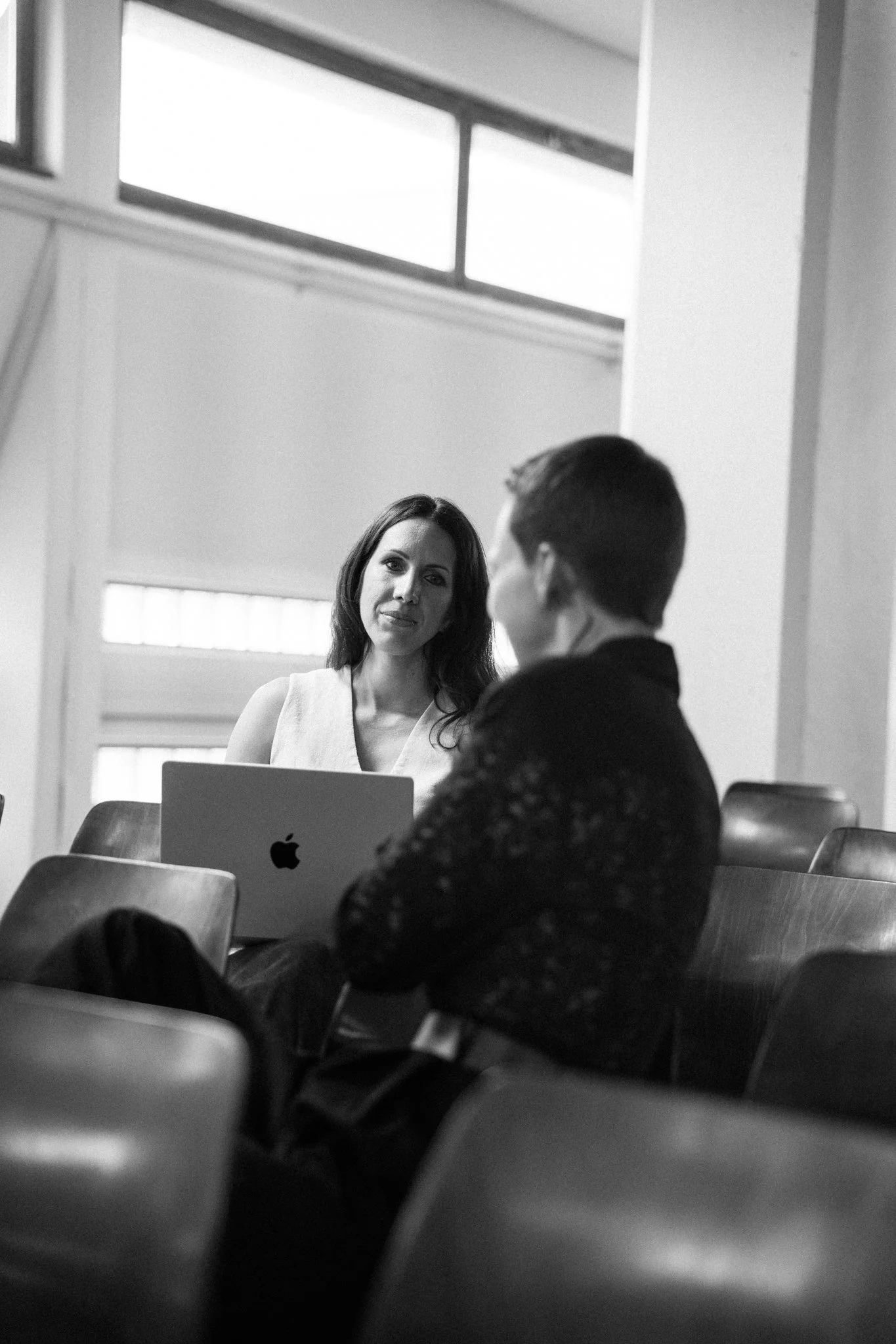Zwei Frauen in einem Raum, eine sitzt an einem Tisch mit einem Laptop, die andere steht und schaut sie an. Es ist eine schwarz-weiß Aufnahme, vermutlich in einem Büroumfeld.