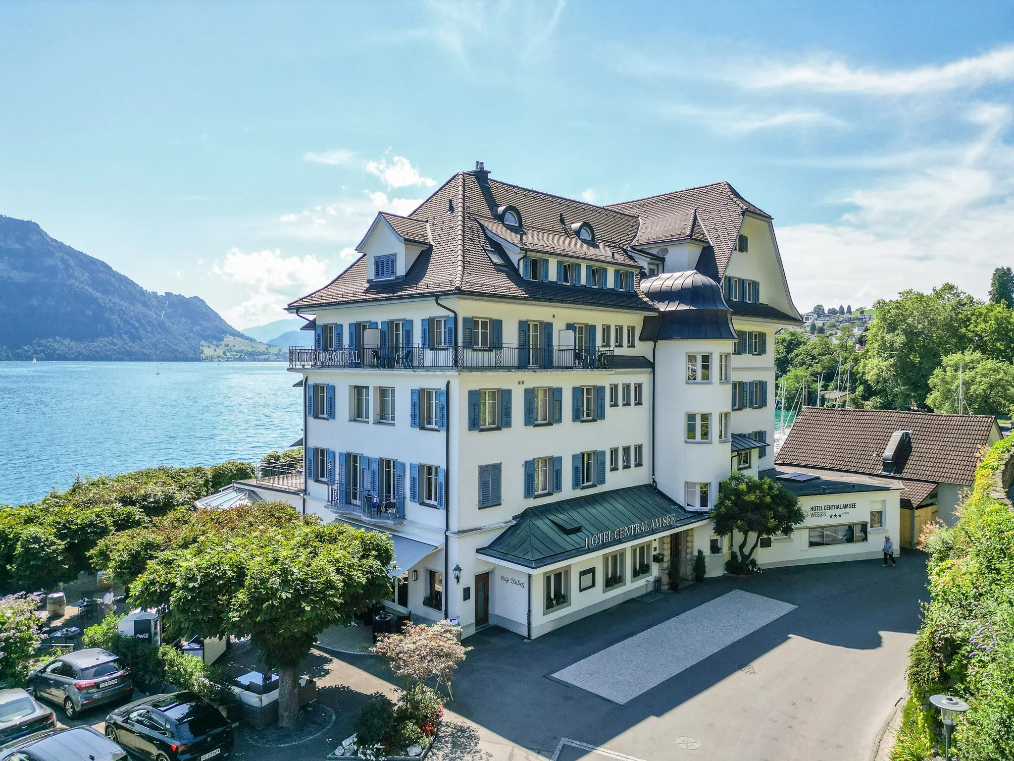 Ein großes, weißes Hotel mit blauen Fensterläden und Dach. Es liegt am Ufer eines Sees mit Bergen im Hintergrund. Es gibt Bäume und einen Parkplatz mit Autos vor dem Hotel.