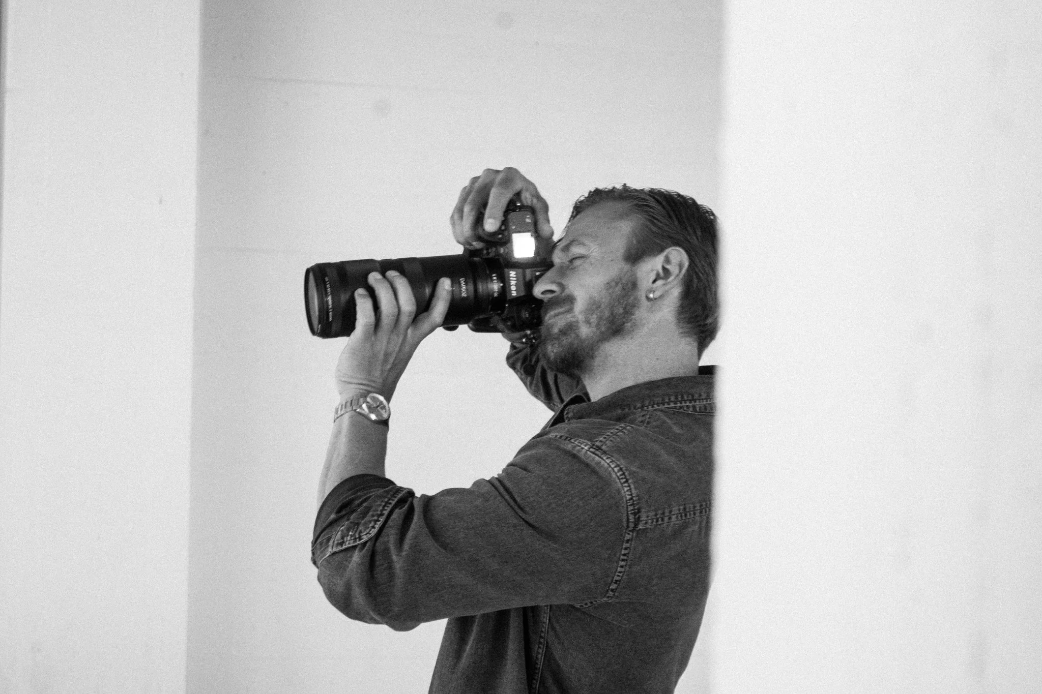 Ein Mann mit dunklem, seitlich gescheiteltem Haar und Earring fotografiert mit einer digitalen Kamera vor einem weißen Hintergrund.