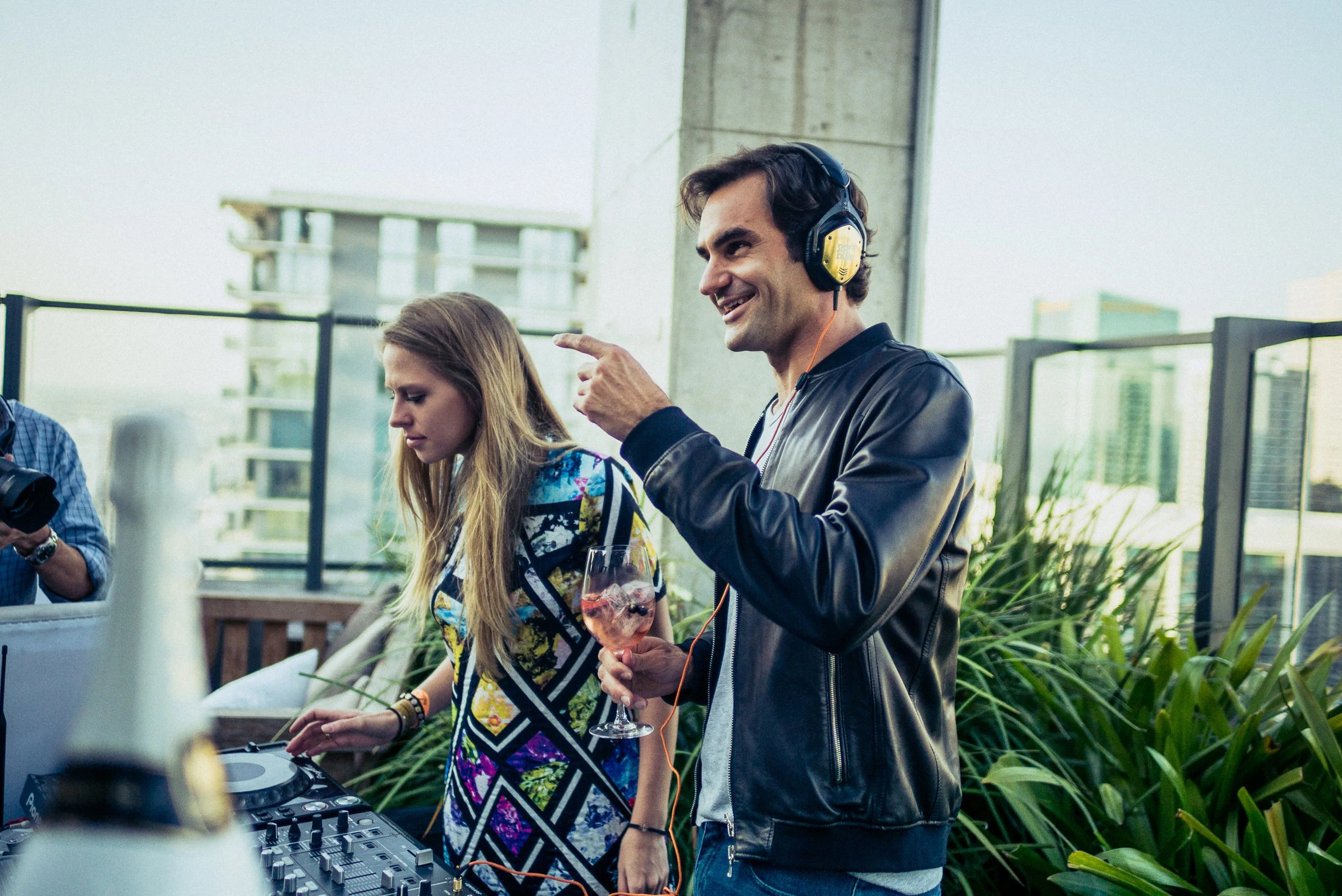 Ein Mann mit dunklen Haaren, schwarzer Lederjacke und gelben Kopfhörern, die er trägt, lächelt und zeigt mit dem Finger, während er an einem DJ-Setup arbeitet. Neben ihm steht eine Frau mit langen, glatten, blonden Haaren, in einem bunten, gemusterte