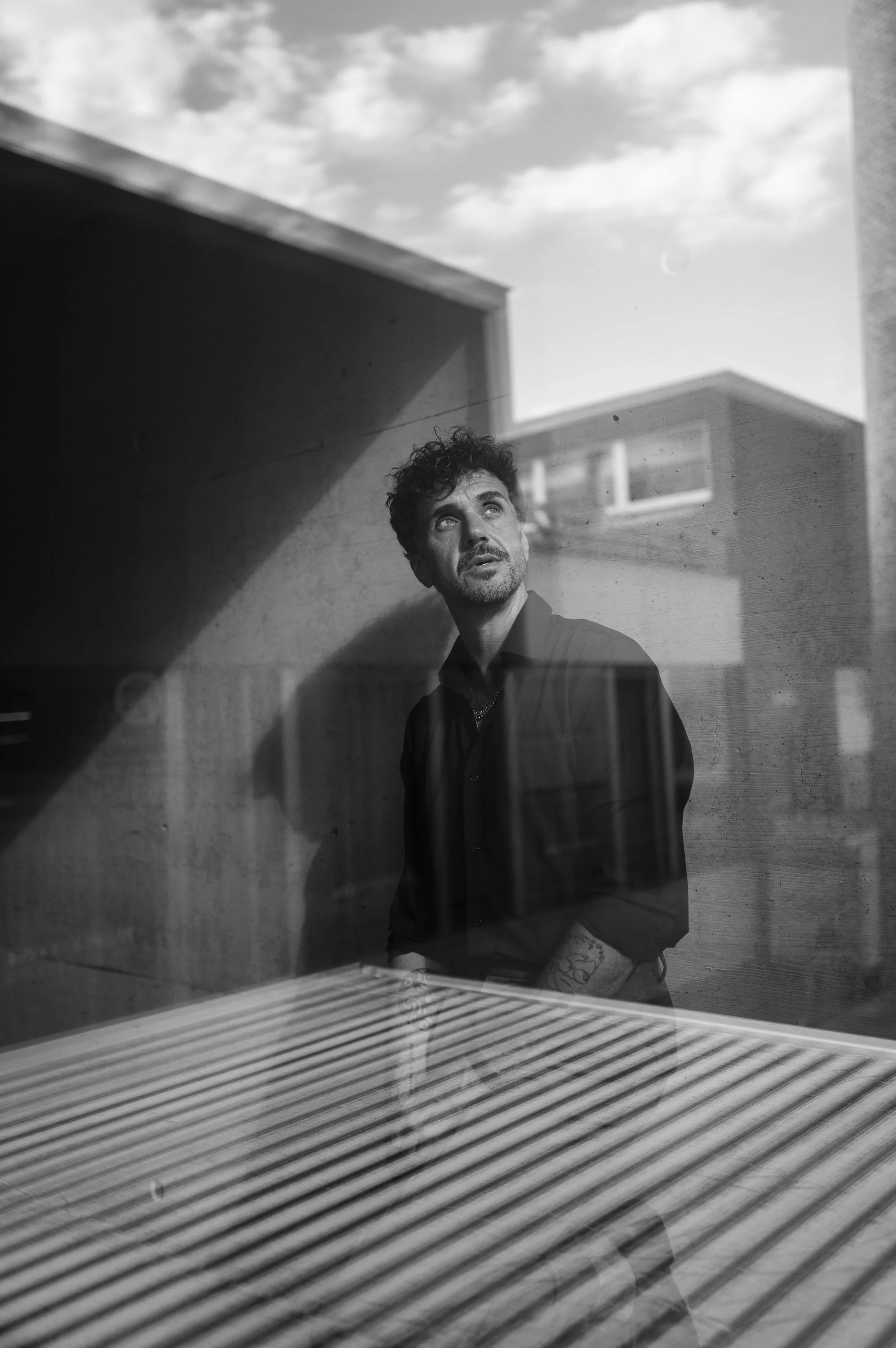Schwarz-weiß-Foto eines Mannes mit curly Haaren, der in einem modernen Gebäude steht und nach oben blickt. Er trägt dunkle Kleidung und hat sichtbare Tattoos am Arm. Das Bild ist durch eine Glasscheibe aufgenommen, die einen Streifeneffekt erzeugt, u