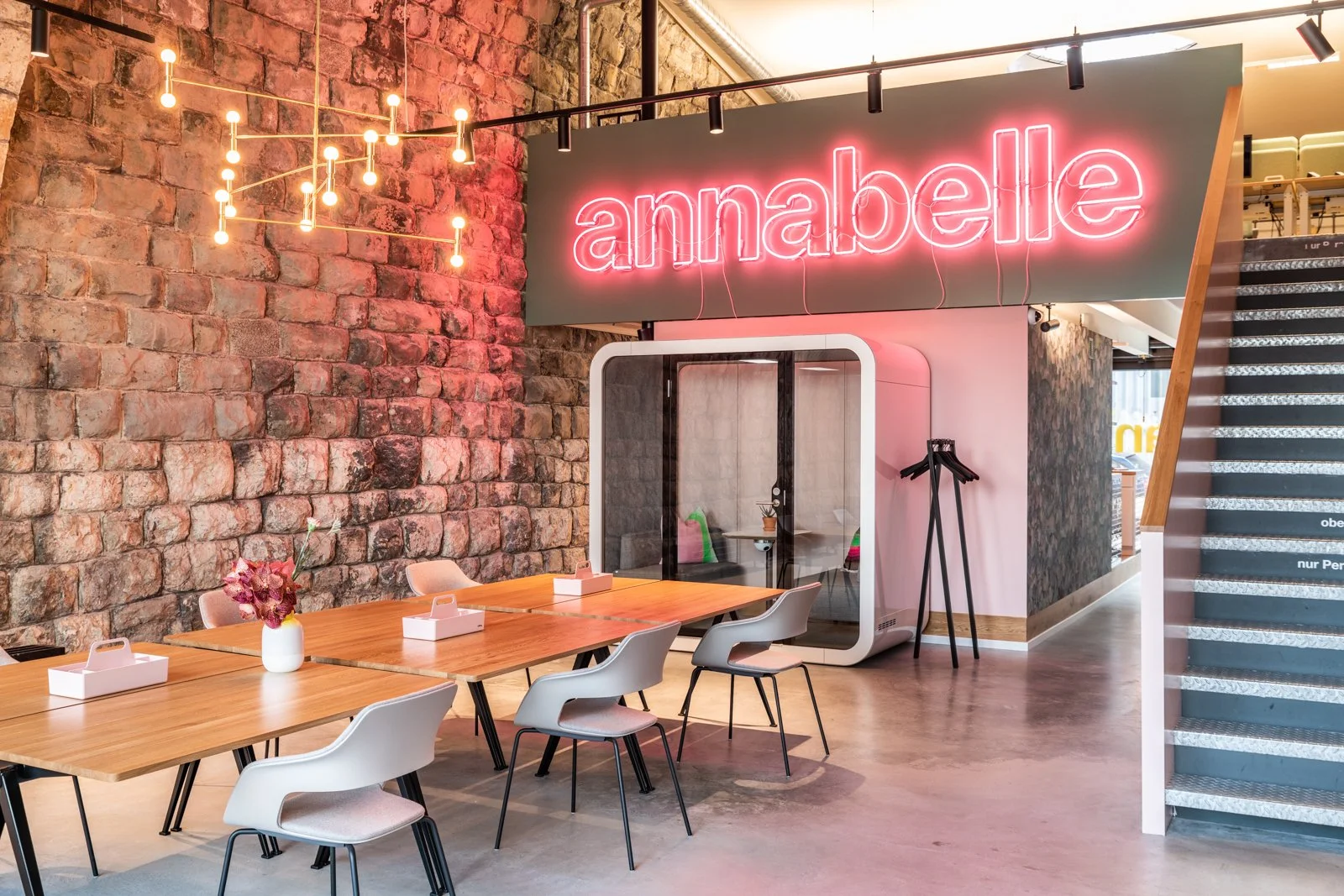Moderner Café-Bereich mit einem großen neonrosa Leuchtschild mit der Aufschrift 'annabelle'. Es gibt einen Holztisch mit weißen Stühlen, eine Vase mit pinken Blumen, einen kleinen Raum für private Gespräche, einen Steharbeitsplatz und eine Treppe auf der rechten Seite. Die Wand besteht aus rohem Ziegelstein und es gibt eine künstlerische Beleuchtung an der Decke.