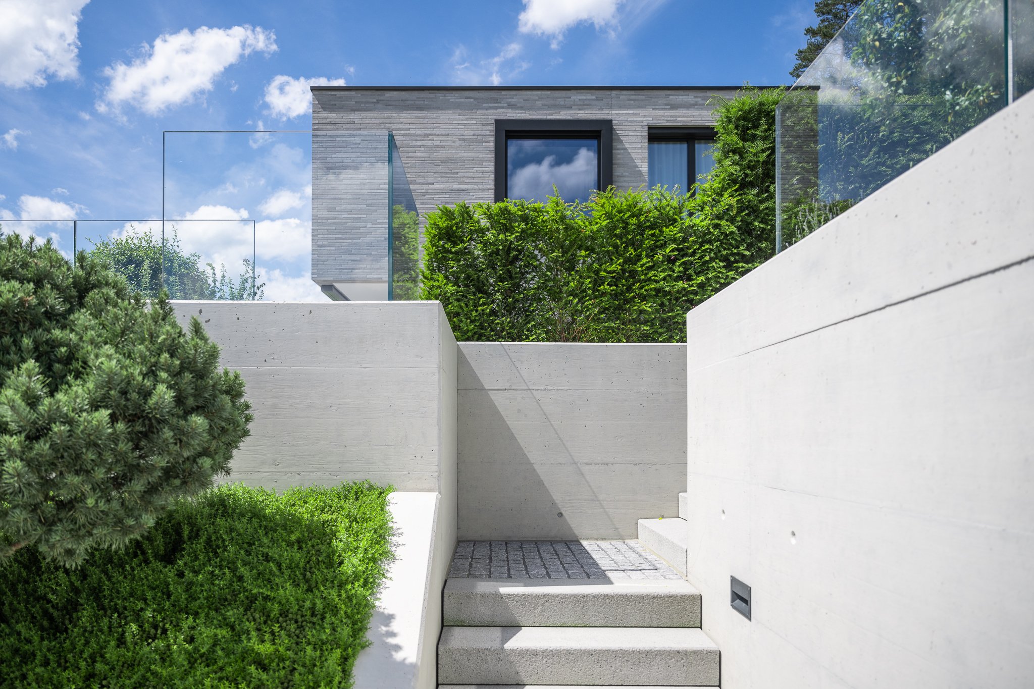 Moderner, minimalistischer Eingangsbereich eines Hauses mit anthrazitfarbenen Fenstern, grünen Büschen und weißen Betonwänden unter blauem Himmel mit Wolken.