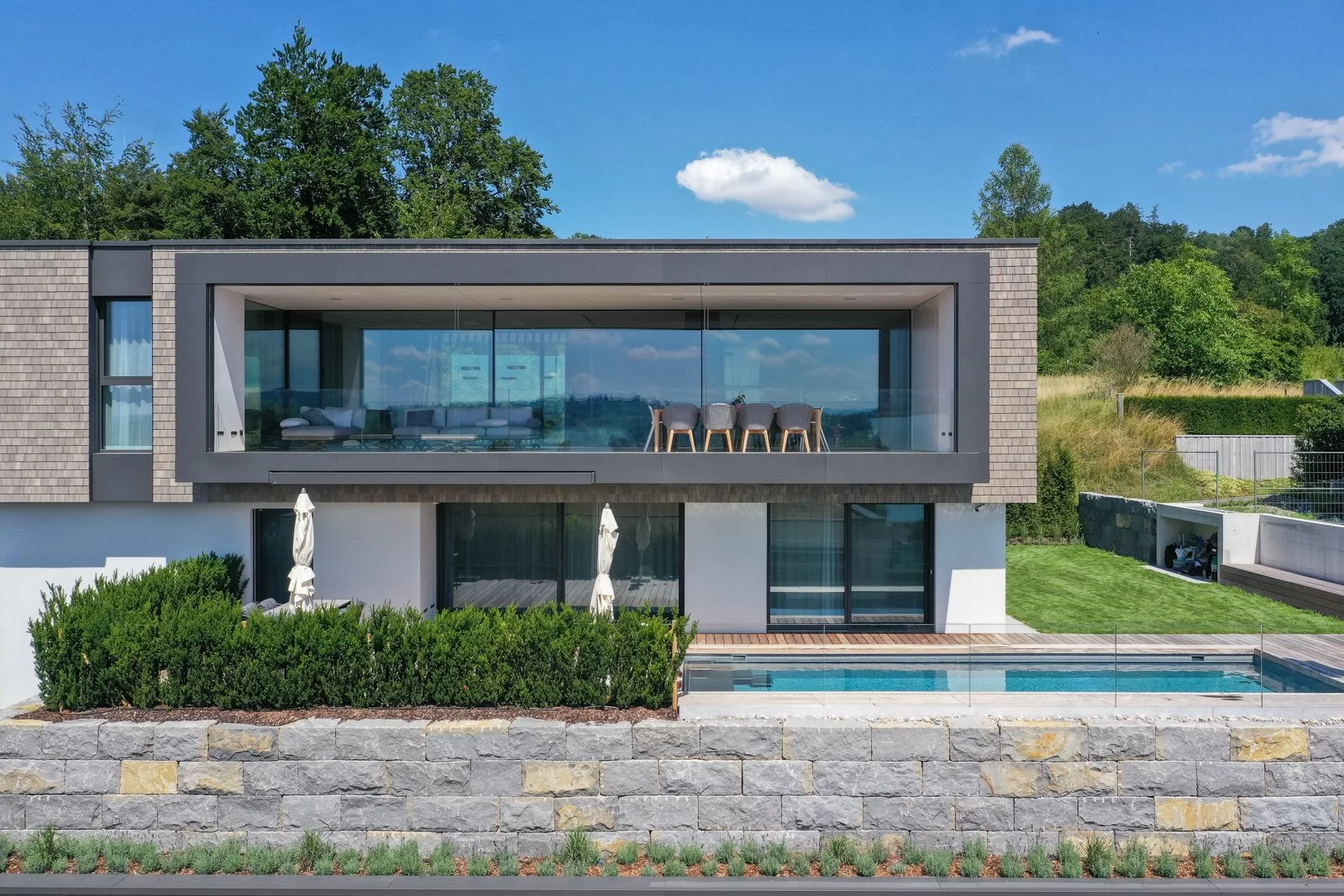 Moderner zweistöckiger Haus mit großen Glasfenstern, Terrasse mit Sitzgruppe, Swimmingpool, umgeben von Grünflächen und Bäumen, bei sonnigem Himmel.