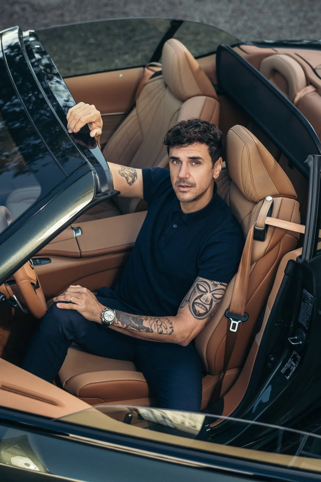 Mann sitzt in einem braunen Cabriolet mit Lederinnenraum, trägt eine schwarze Polohemd und hat Tattoos auf den Armen.