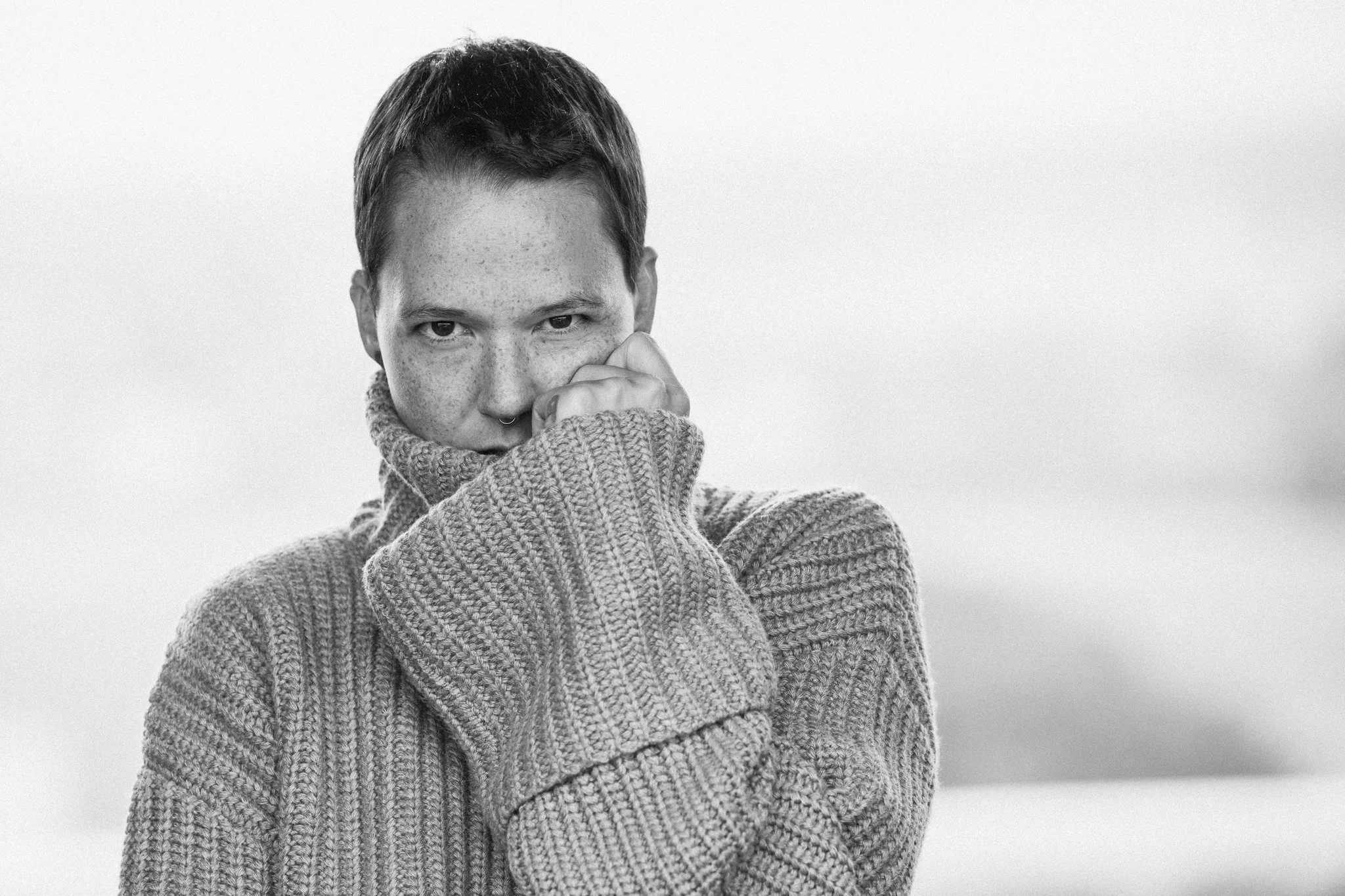 Schwarz-weiß-Foto einer Person mit kurzen Haaren, die einen grauen Pullover trägt, die Hand vor dem Mund hält und in die Kamera schaut.