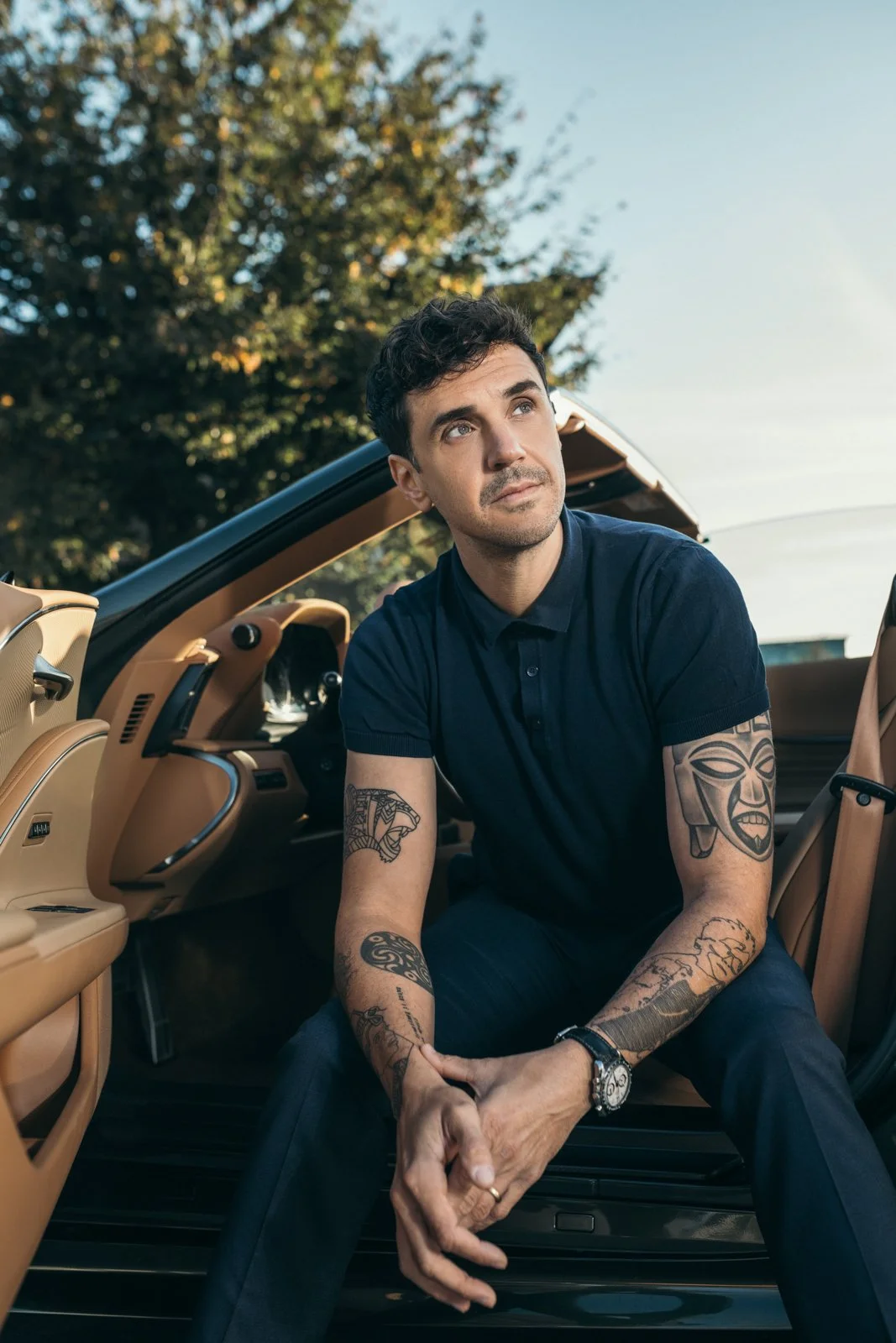 Ein junger Mann mit Tattoos sitzt in einem offenen Auto, schaut nach oben, bei Tageslicht, mit Bäumen im Hintergrund.