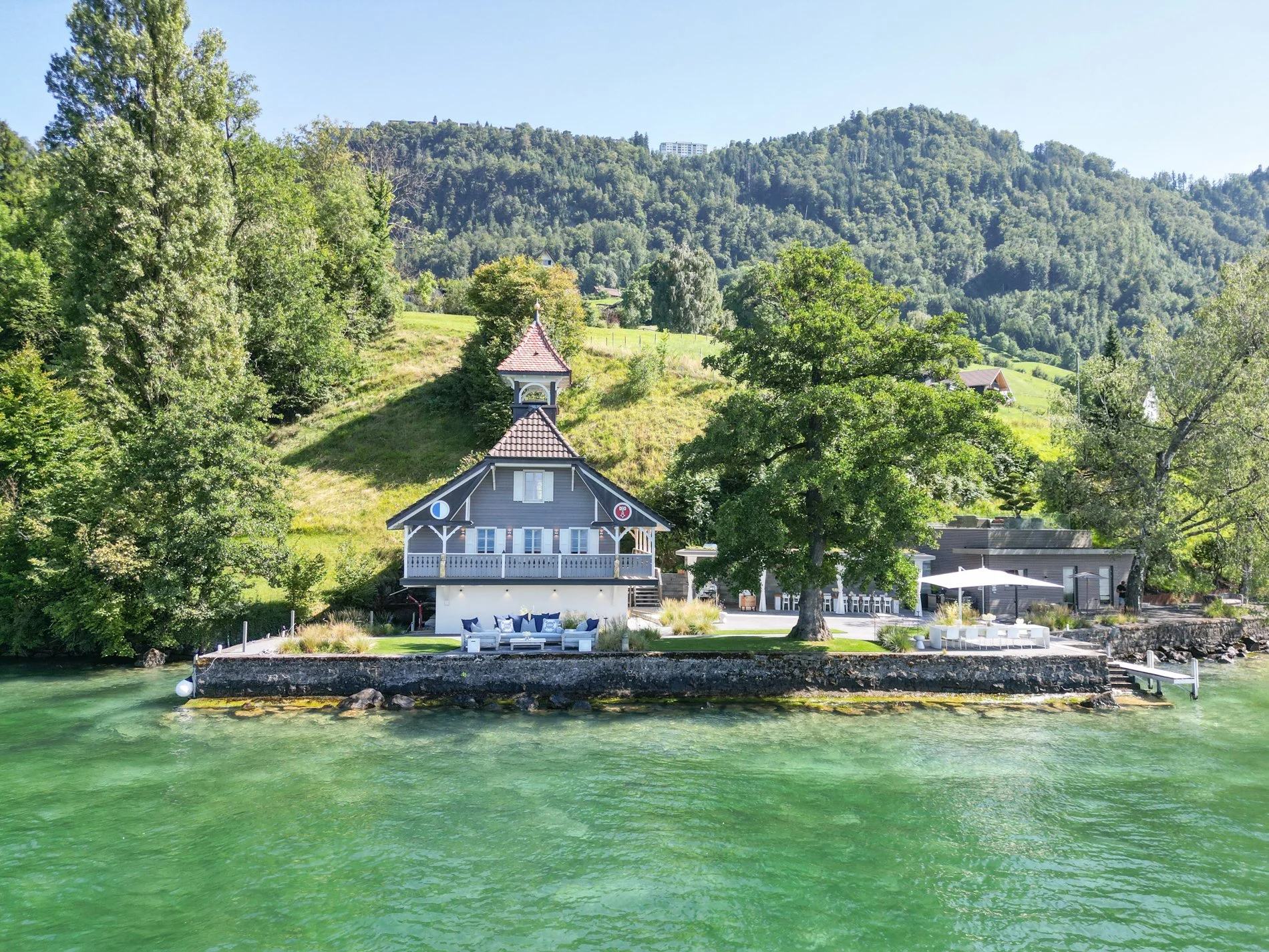 Ein Haus am Wasser mit einer Terrasse, umgeben von Bäumen, im Hintergrund grüne Hügel und ein blauer Himmel.