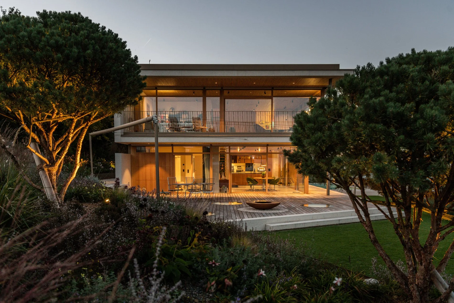 Moderne zweistöckige Villa bei Sonnenuntergang, umgeben von Bäumen und Garten, mit offenem Wohnbereich im Erdgeschoss und Balkon im Obergeschoss, stilvoll eingerichtet.