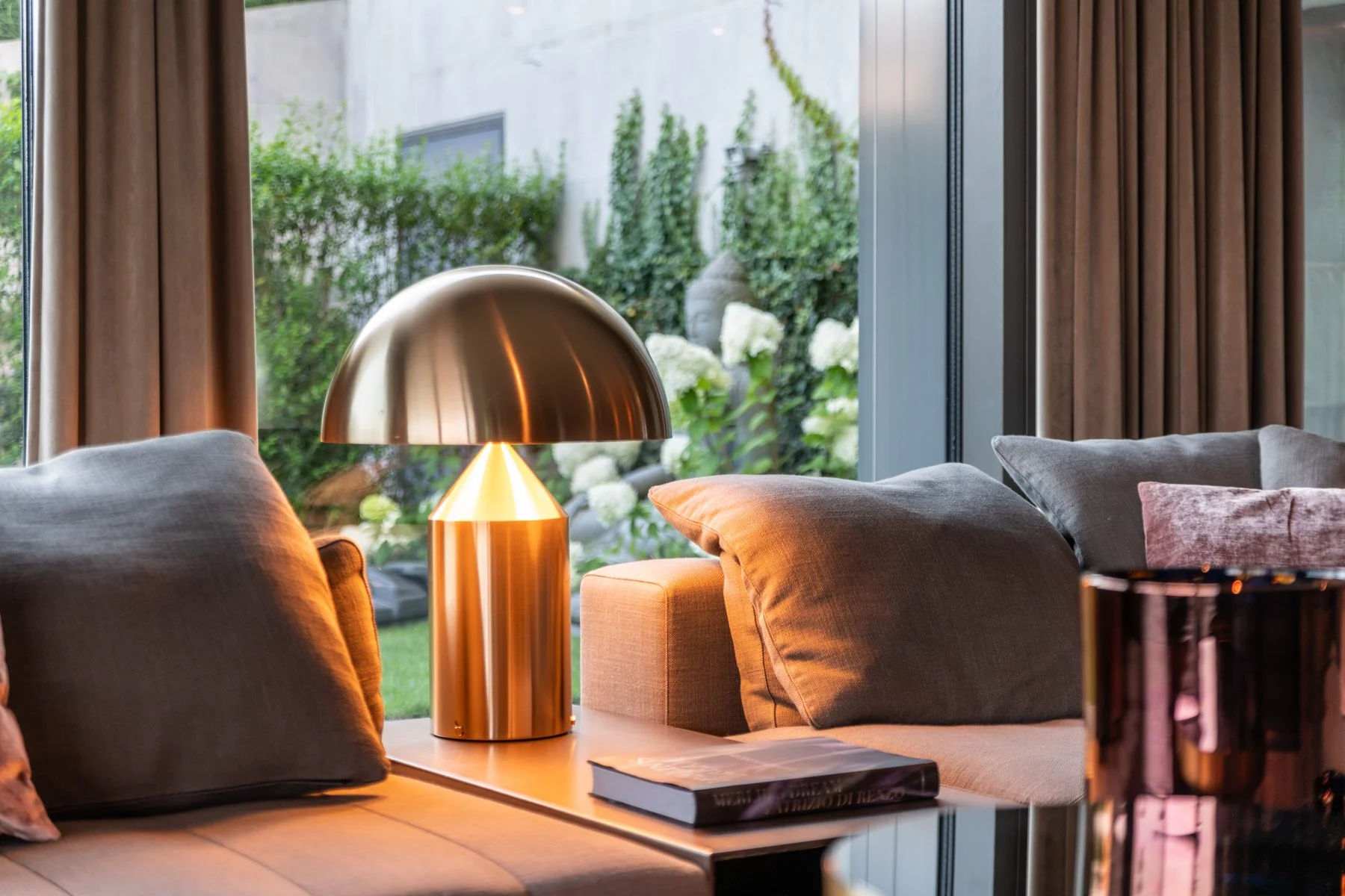 Innenaufnahme eines Wohnzimmers mit grauen Sofas, braunen Vorhängen, einem goldfarbenen Tischlampe und einem Fenster mit Blick auf einen Garten mit weißen Blumen und grünen Sträuchern.