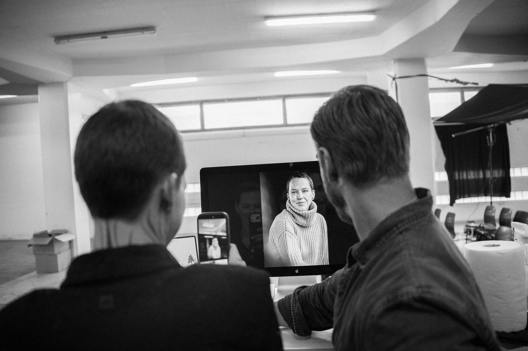 Drei Personen beobachten eine Person auf einem Computerbildschirm, die eine Frau in einem grauen Pullover zeigt, während im Hintergrund große Fenster den Raum beleuchten.