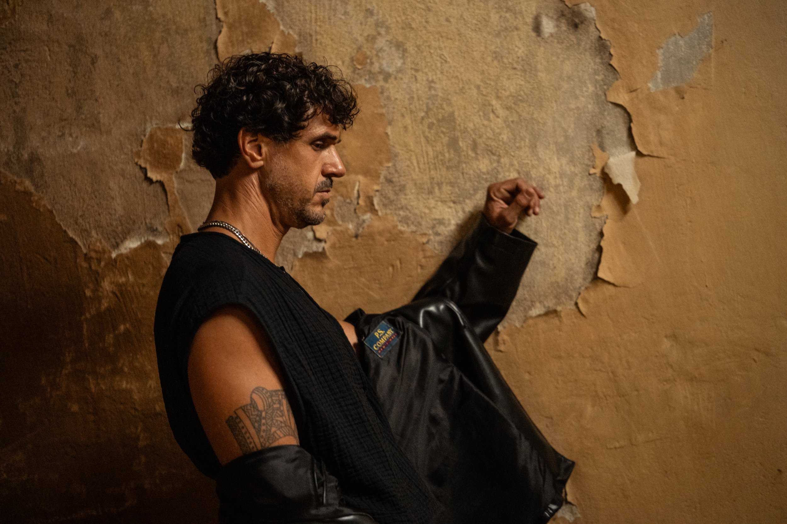 Ein Mann mit dunklen, lockigen Haaren, dunkler Haut, Tattoos am Arm, trägt schwarze Kleidung und einen silbernen Kettenhals, steht vor einer alten, zerfallenden Wand.