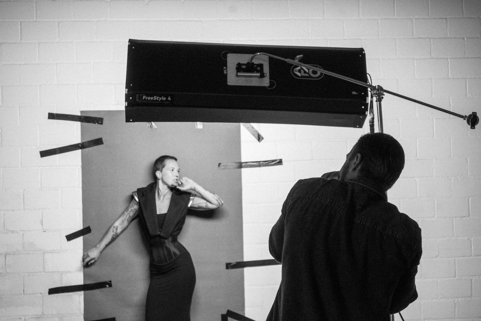 Ein Fotograf fotografiert eine tattoosierte Frau, die vor einer Wand mit Spiegeln und Klebetapes steht.