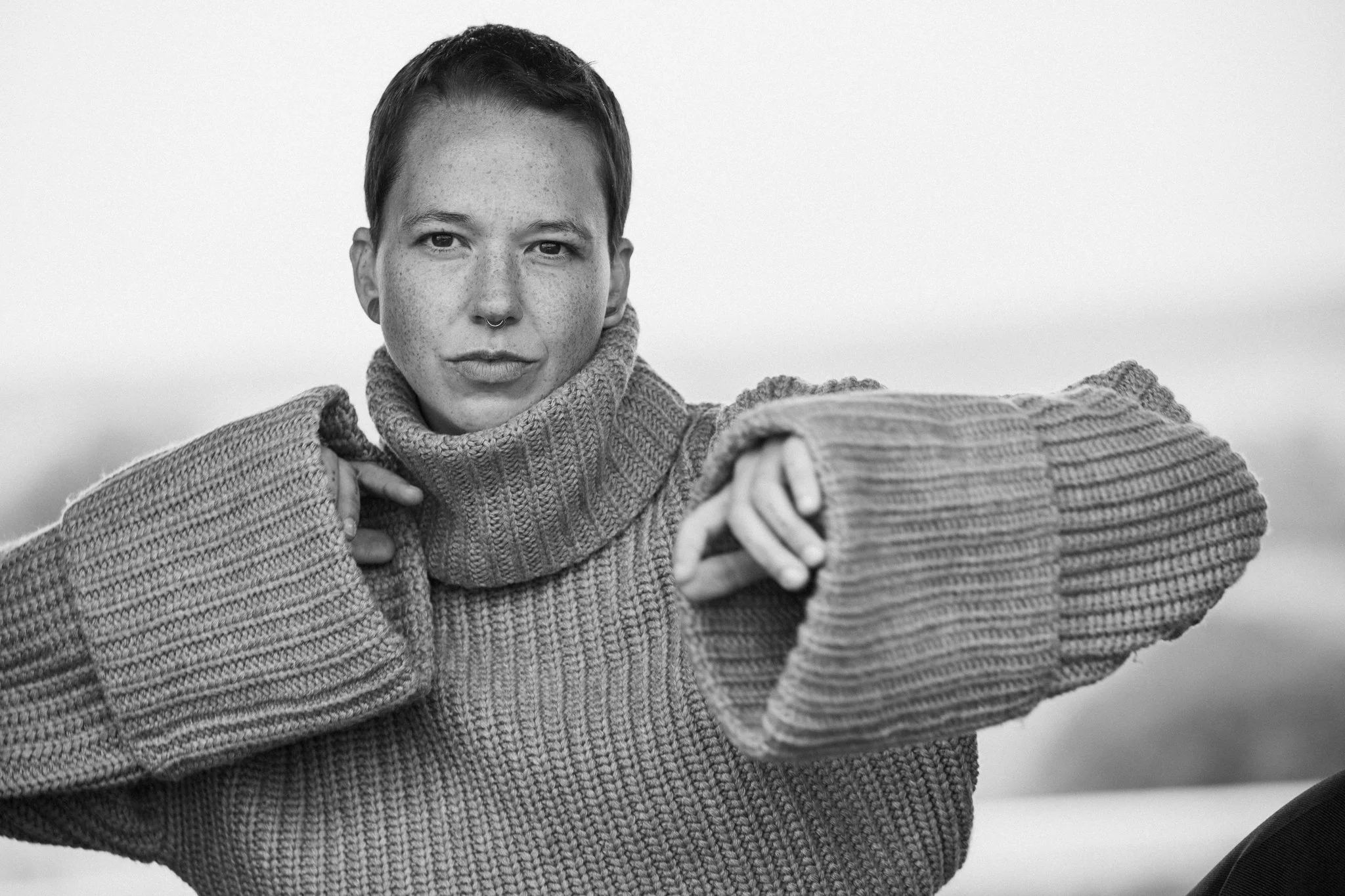 Schwarz-weiß-Bild eines jungen Menschen mit kurzen Haaren, der eine große Strickjacke trägt und in die Kamera schaut.