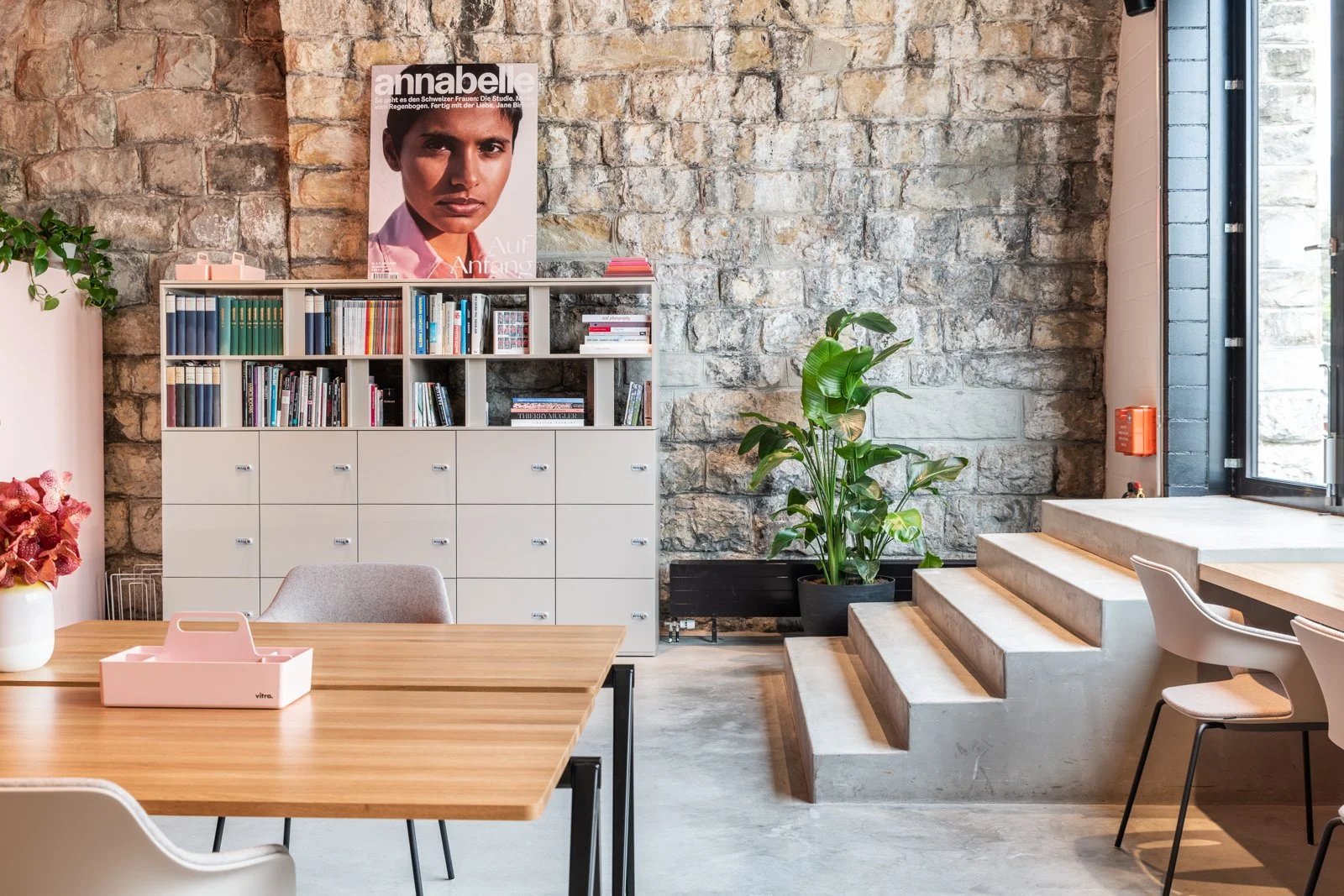 Ein modernes Büro mit einem Holztisch, weißen Stühlen, einer weißen Bücherregal mit Büchern, einer Pflanze, einem Bücherregal und einer großen Wand mit sichtbaren Ziegelsteinen. Es gibt auch Stufen, die zu einem erhöhten Bereich führen, und große Fenster, die natürliches Licht hereinlassen.