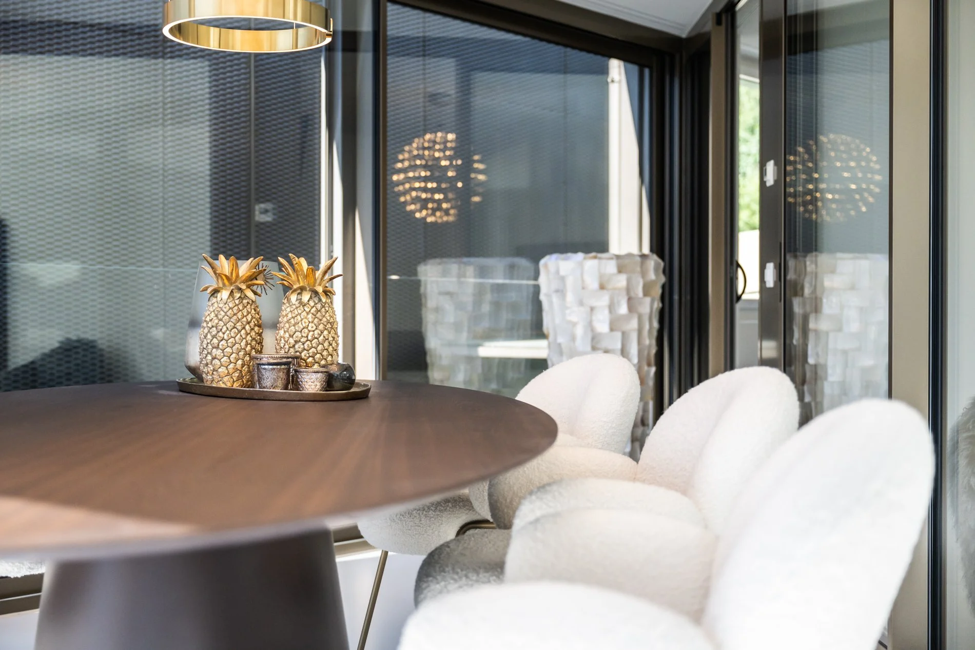 Ein moderner Essbereich mit einem runden Holztisch und weißen, gepolsterten Stühlen, Dekorationsvase mit Ananas-Design auf dem Tisch, große Glasflächen mit schwarzen Rahmen, reflektierende Pendelleuchten, und einer weißen Keramik-Skulptur im Hintergrund.