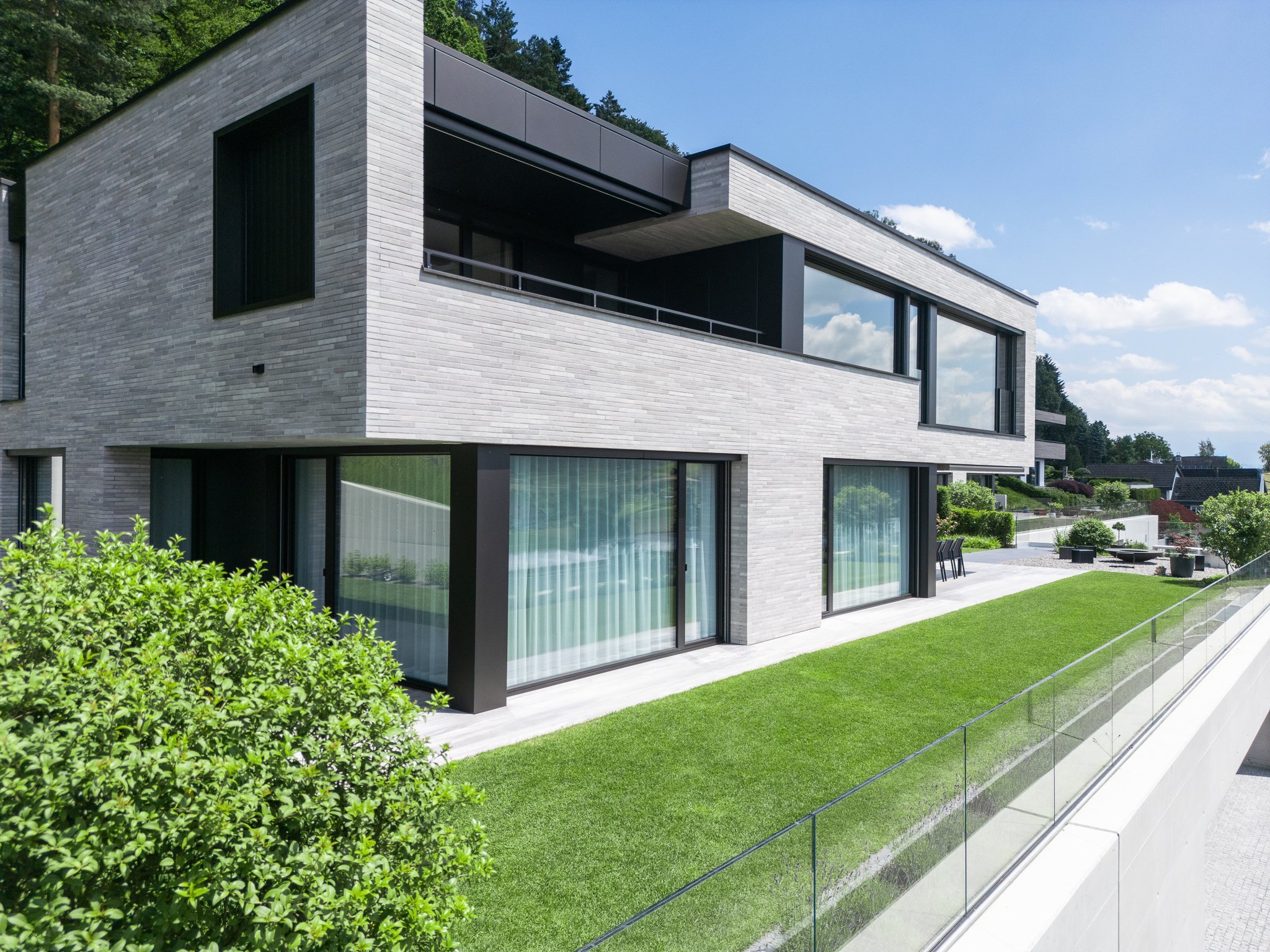 Moderne, mehrstöckige Villa mit großen Glasfenstern, grauen Ziegelwänden und einem Balkon, umgeben von Grünland mit Büschen, Bäumen und einem klaren blauen Himmel.