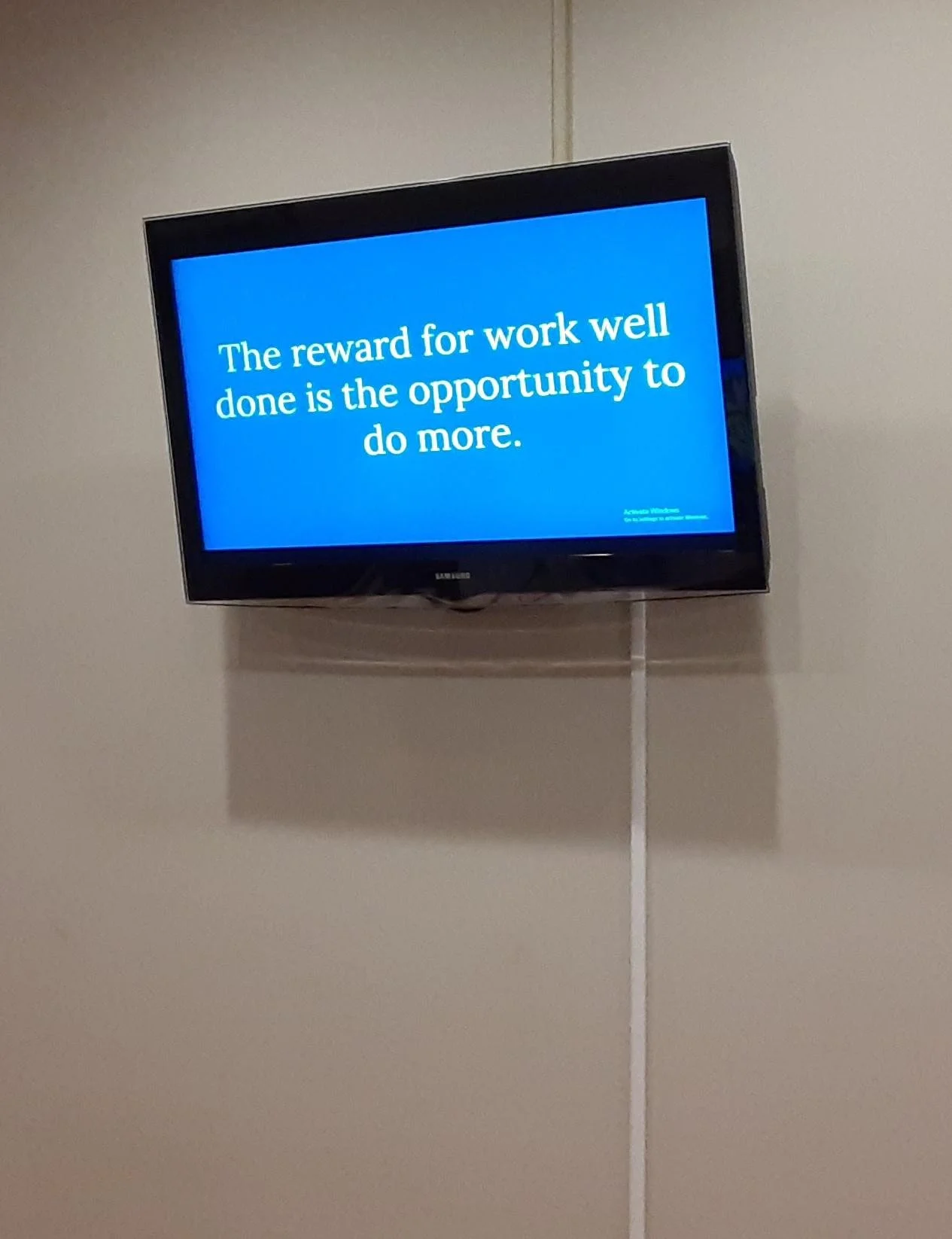 A flat-screen TV mounted on a wall displaying a blue screen with white text that reads, "The reward for work well done is the opportunity to do more."