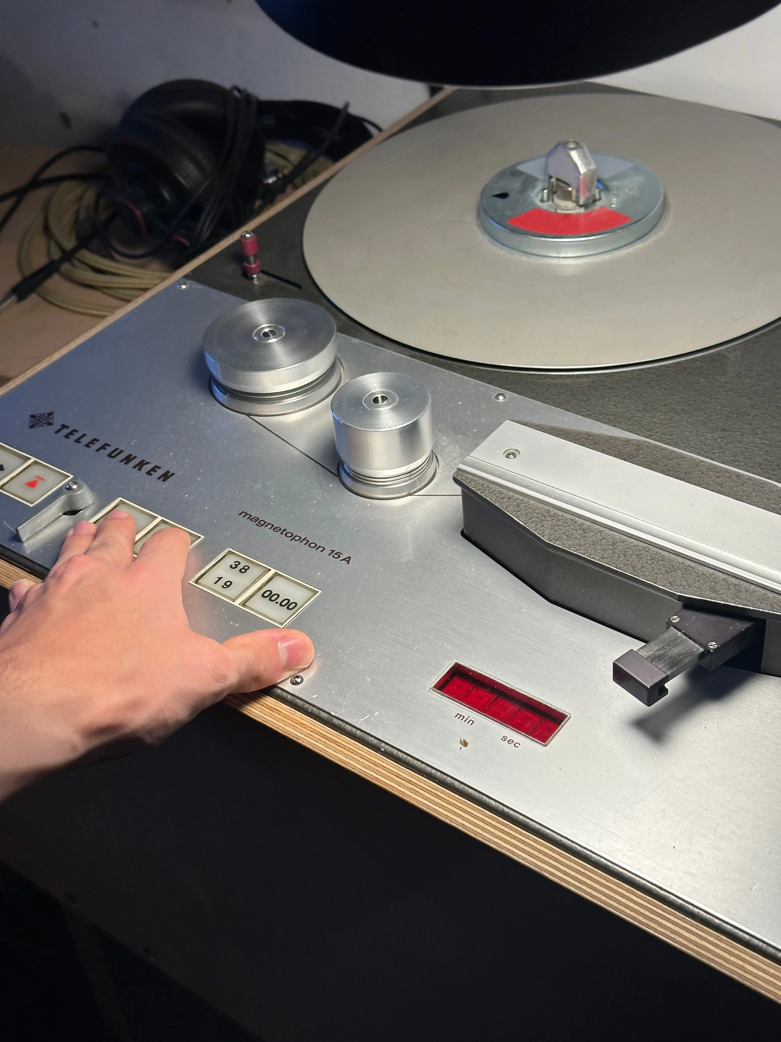 Close-up of a vintage Telefunken reel-to-reel tape recorder with metallic knobs, buttons, and a needle indicator, and part of a person's hand pressing a button.