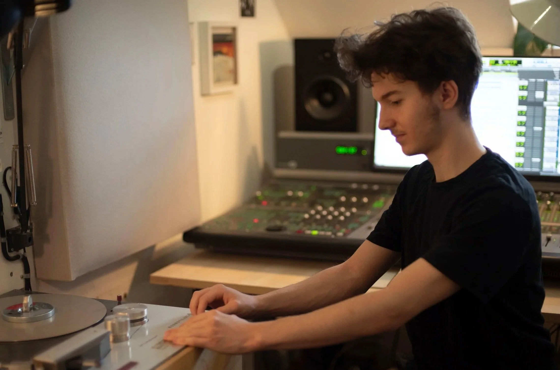 A young man with curly dark hair wearing a black shirt sitting at a mixing console in a recording studio, with studio monitors and computer screens in the background.