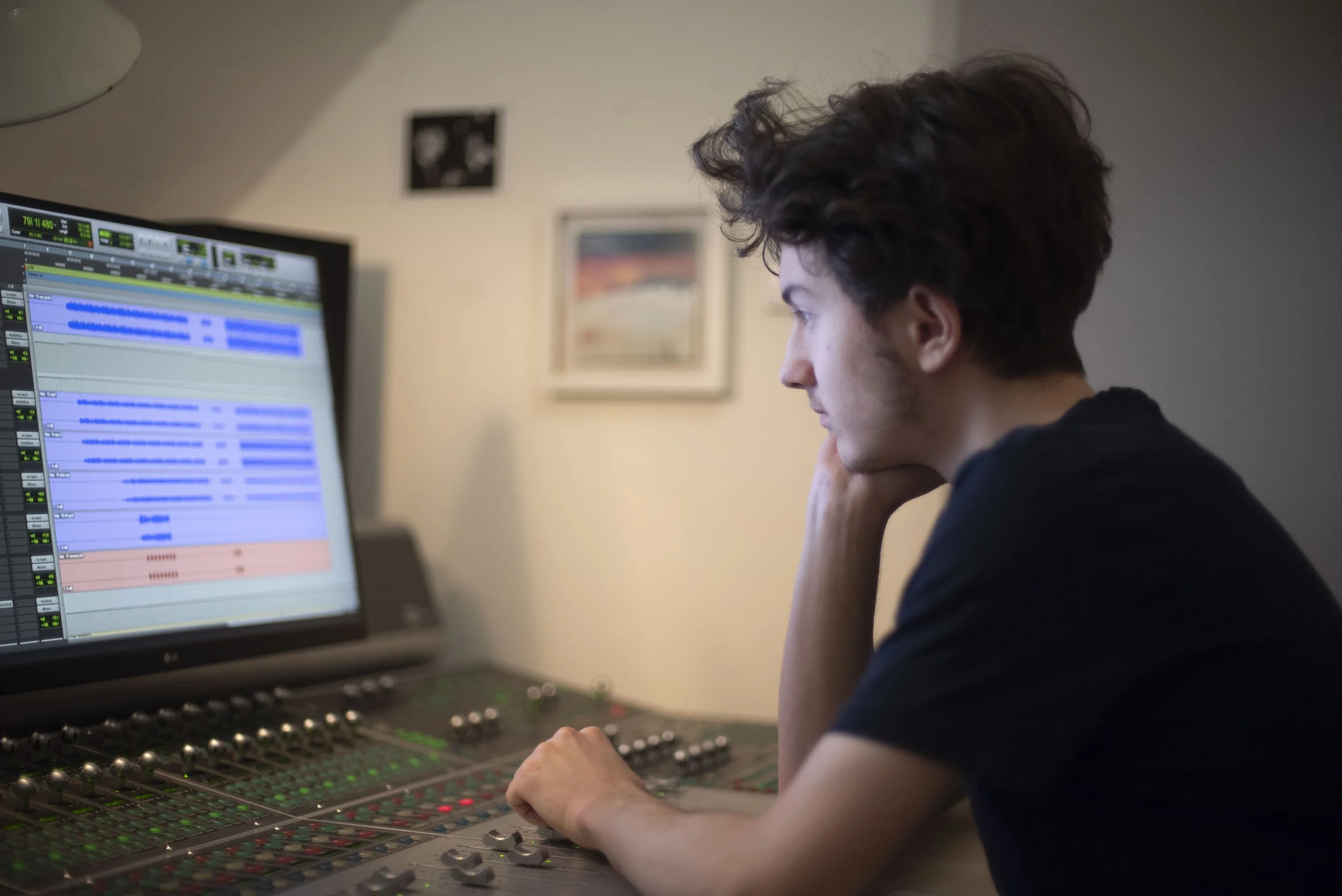 A person with dark curly hair sitting at a mixing console in front of a computer monitor with audio editing software.