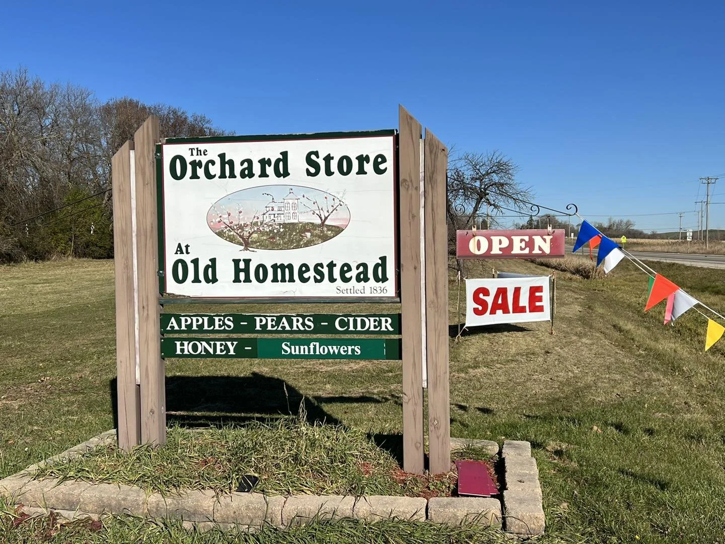 A roadside sign advertising The Orchard Store at Old Homestead, offering apples, pears, cider, honey, and sunflowers. The sign also features an 'Open' sign and a 'Sale' sign, with a blue sky, some trees, and power lines in the background.