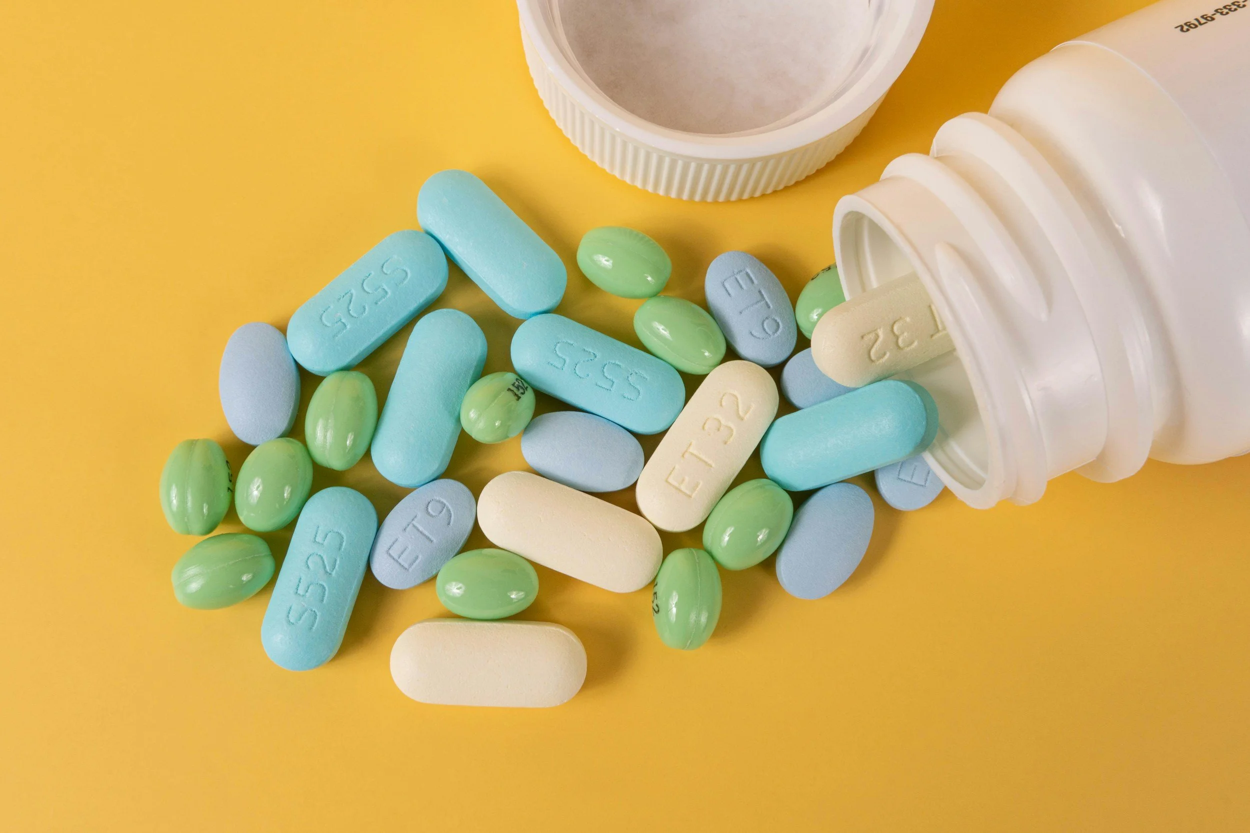 An open prescription pill bottle spilling out various blue, green, and white pills onto a yellow background.