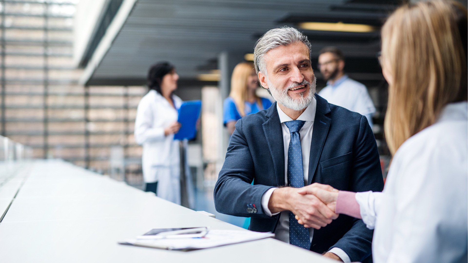 A professional man with gray hair and a beard, dressed in a dark suit and blue tie, smiling as he shakes hands with a woman in a white blazer during a business meeting in a modern office space.