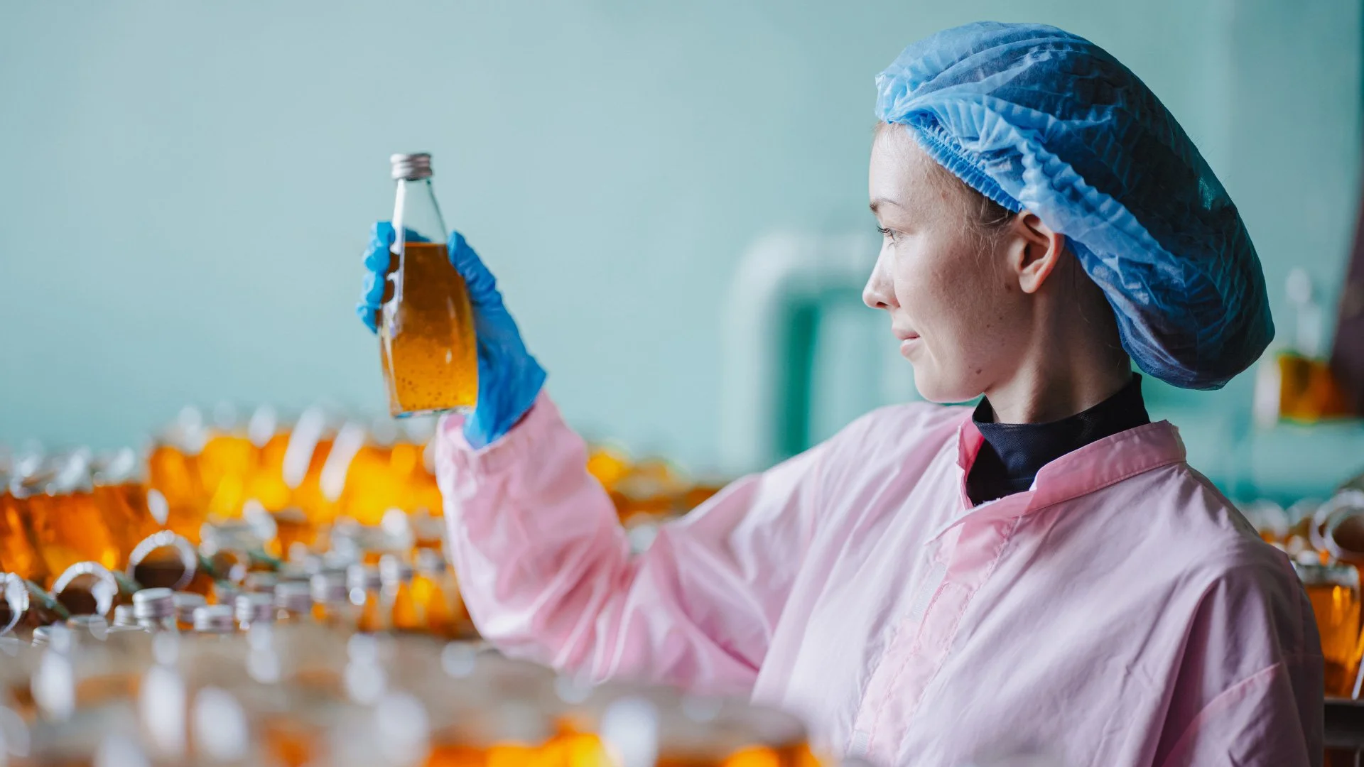A woman in a pink lab coat, blue hairnet, and gloves inspecting a small bottle of amber liquid in a laboratory setting with multiple similar bottles in the background.