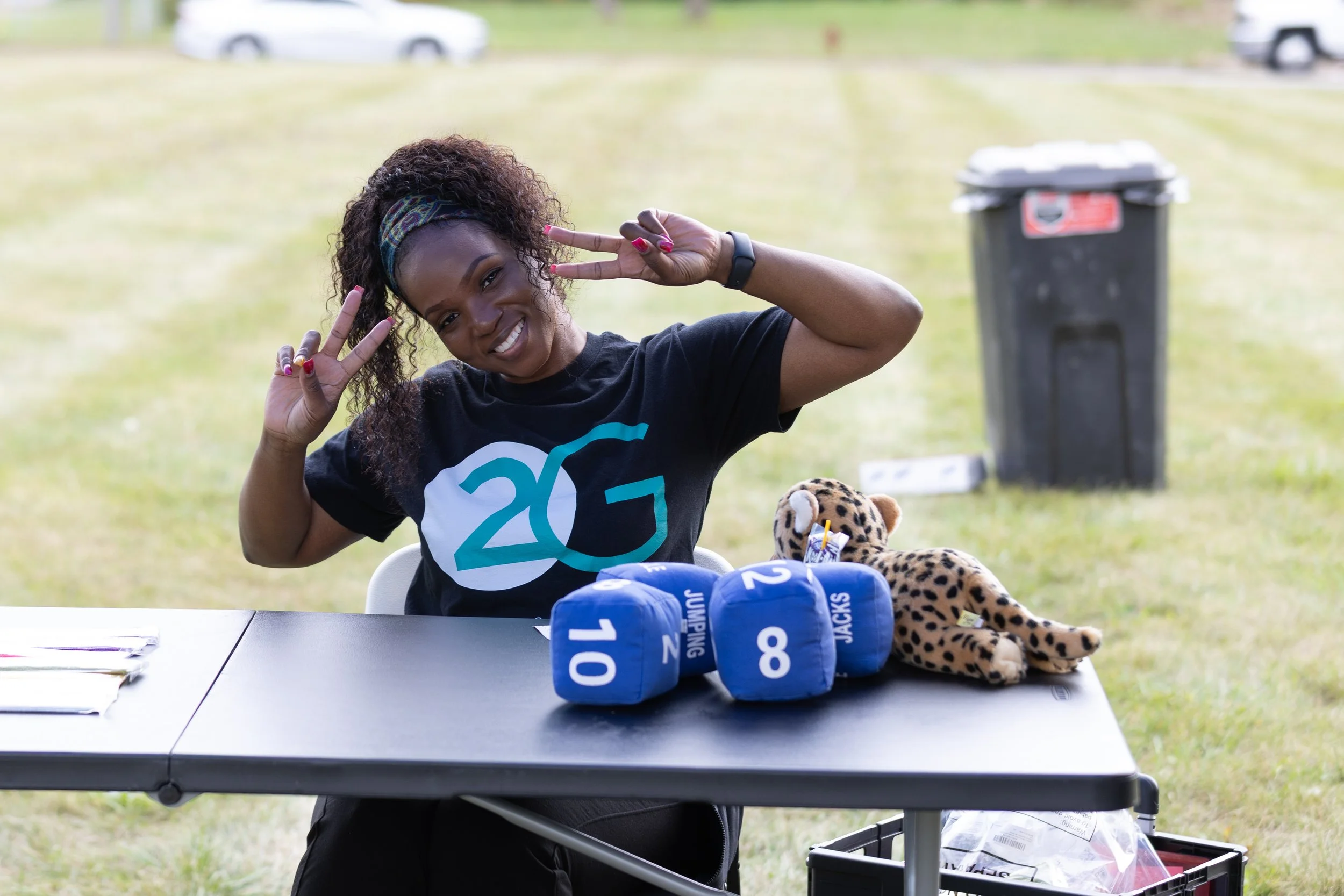 A woman smiling and making peace signs with both hands at an outdoor event, seated at a table with blue foam number blocks and a stuffed leopard toy, with a trash bin and parked cars in the background.