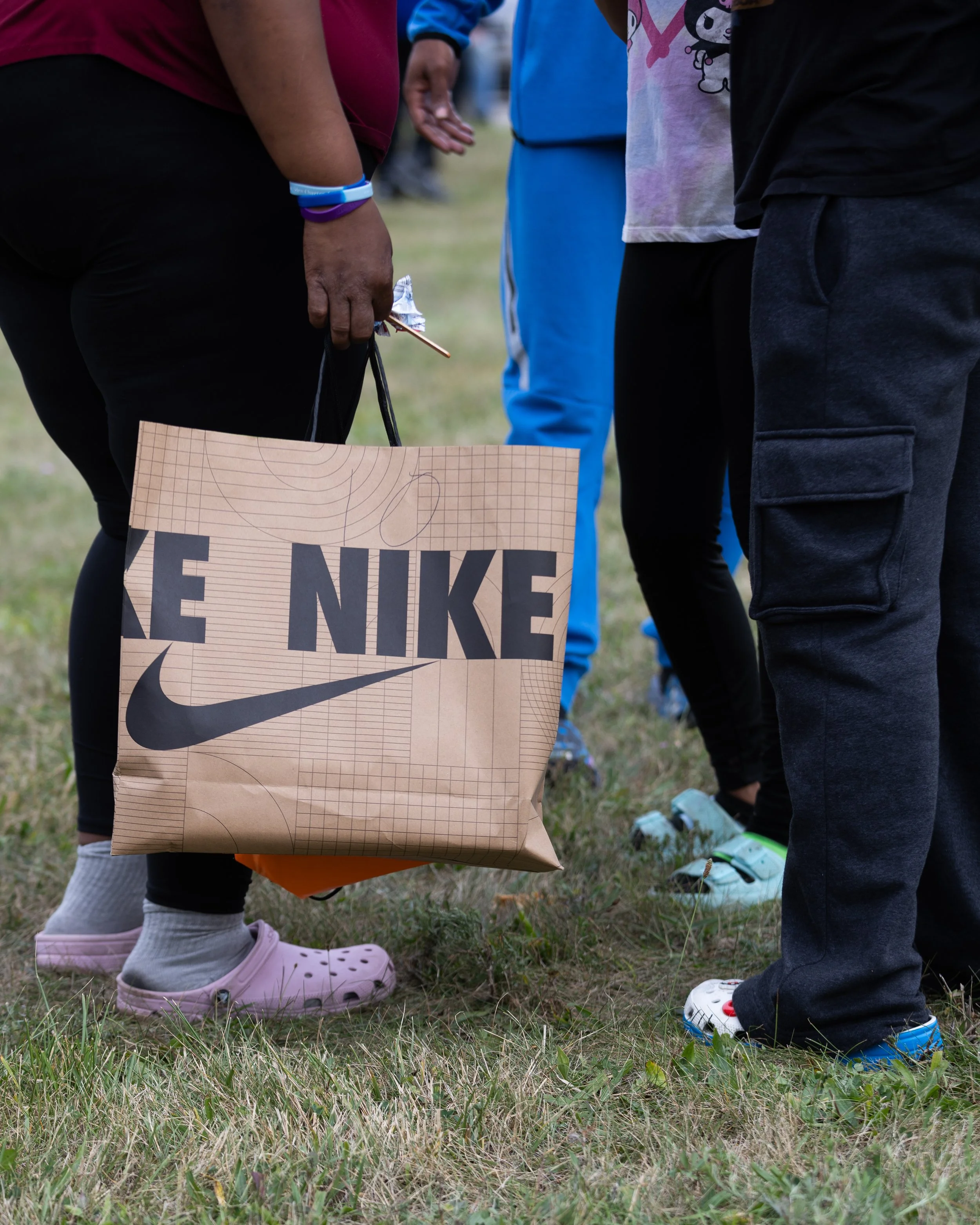 Close-up of children standing outdoors on grass, wearing sneakers and casual clothing, with a brown paper bag and people in the background.