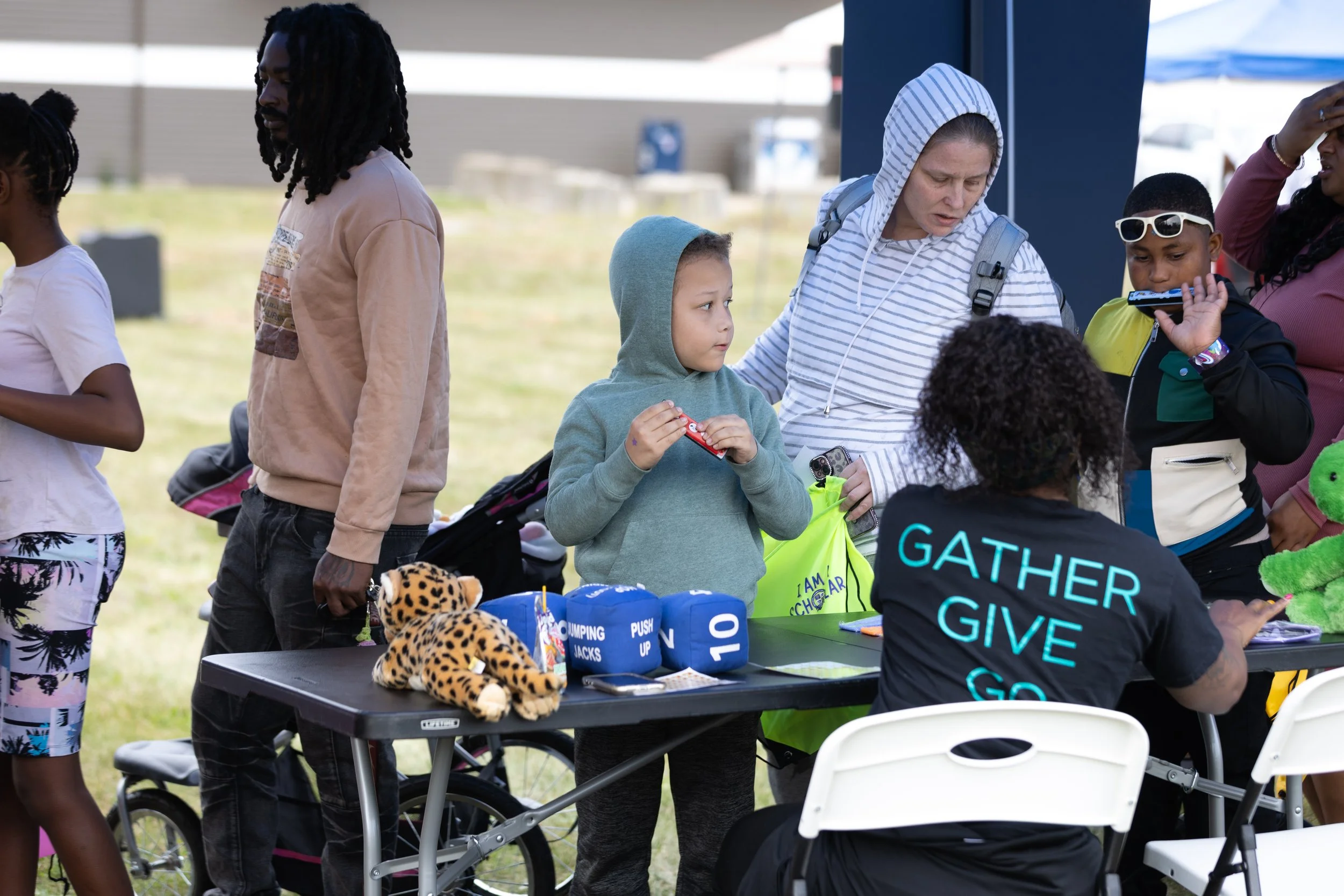 People gathering at an outdoor event with a table displaying prizes and toys, including stuffed animals, under a tent on a grassy area.