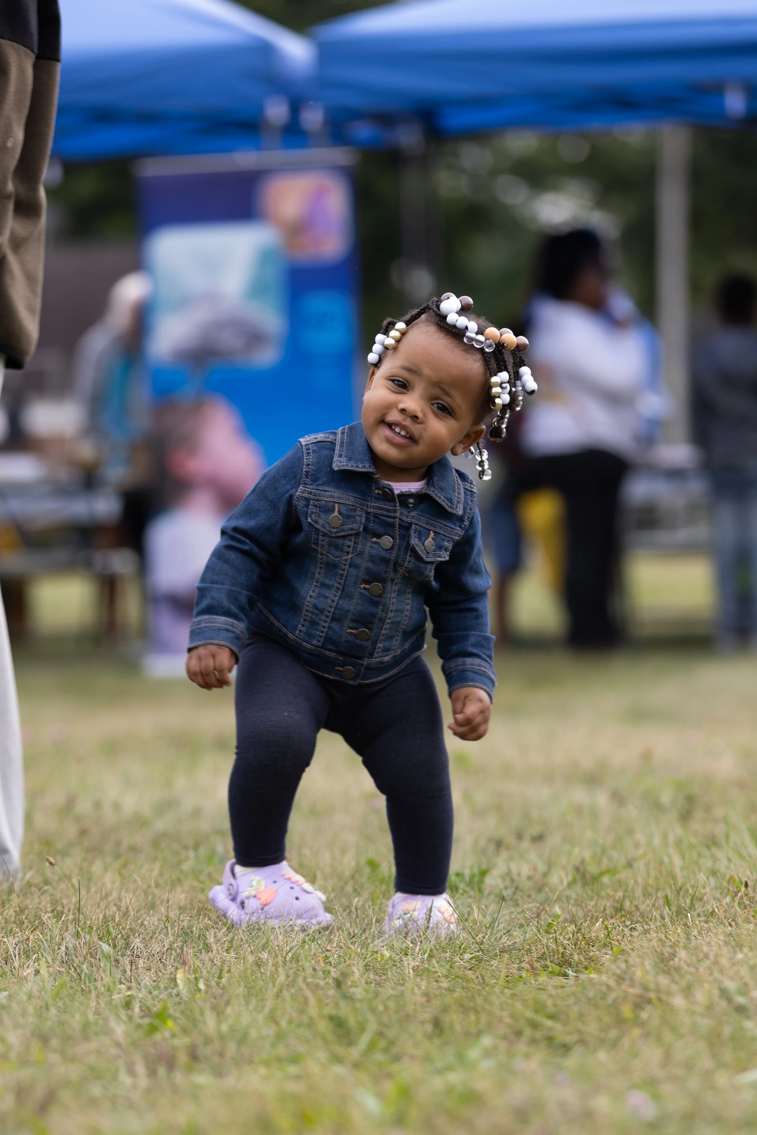 Young girl with bead braids and denim jacket standing on grass at outdoor event, smiling and squatting, with people and tents in the background.