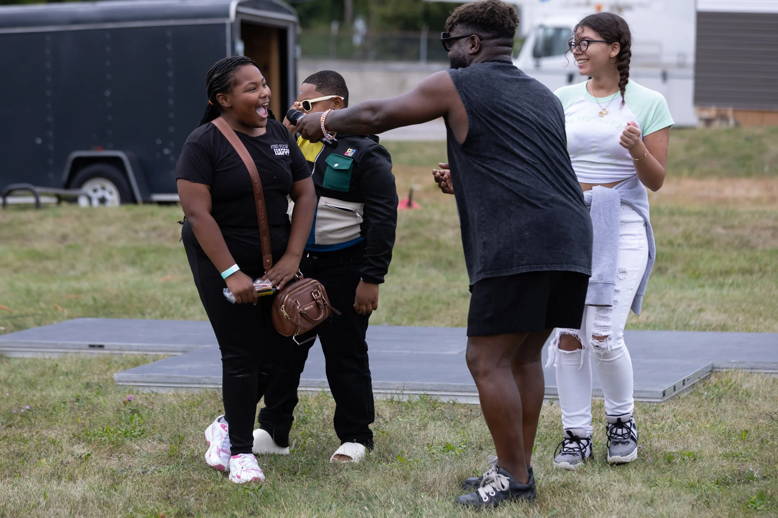 A group of four young people, consisting of two girls and two boys, standing outdoors on grass in front of a trailer and van, participating in a fun activity involving a microphone and a platform, smiling and interacting with each other.