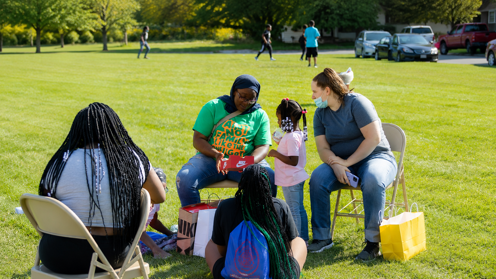 A group of five women and two young girls sitting on chairs and on the grass in a park during daytime, with trees, parked cars, and a few people walking in the background. They are engaged in a conversation, with some holding shopping bags and one woman wearing a face mask.
