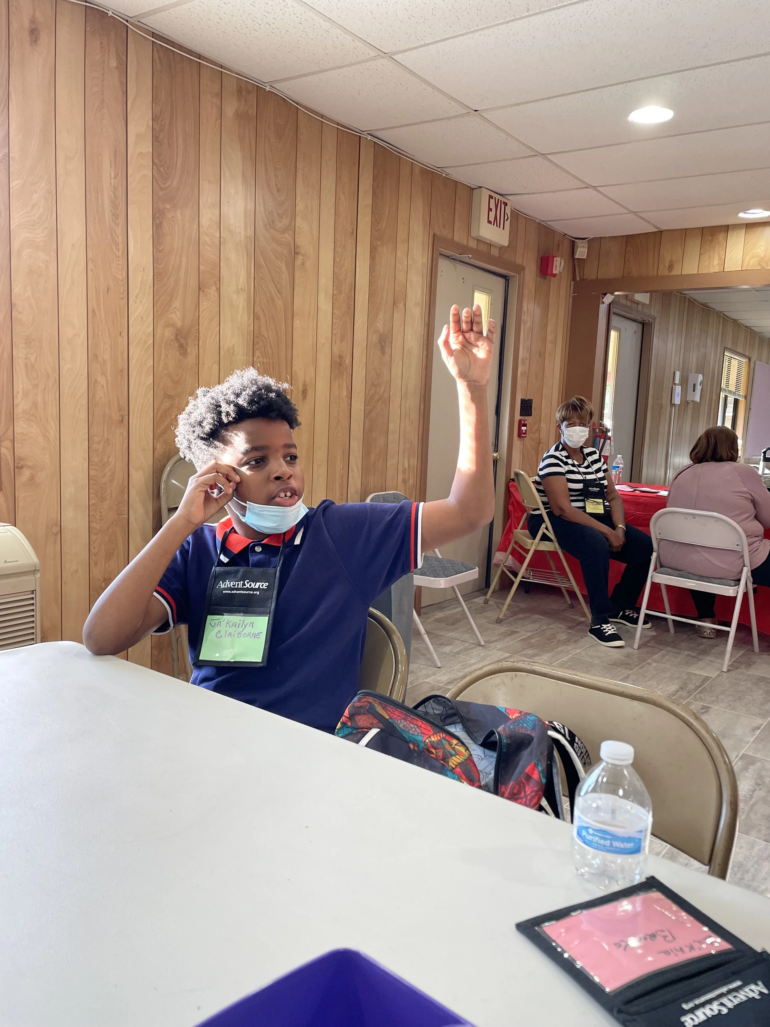 A young boy with curly hair and a face mask raising his hand in a room with wood paneling, a woman with short hair and a face mask sitting at a table in the background, and tables with chairs. There is a water bottle and a pink folder on the table in the foreground.