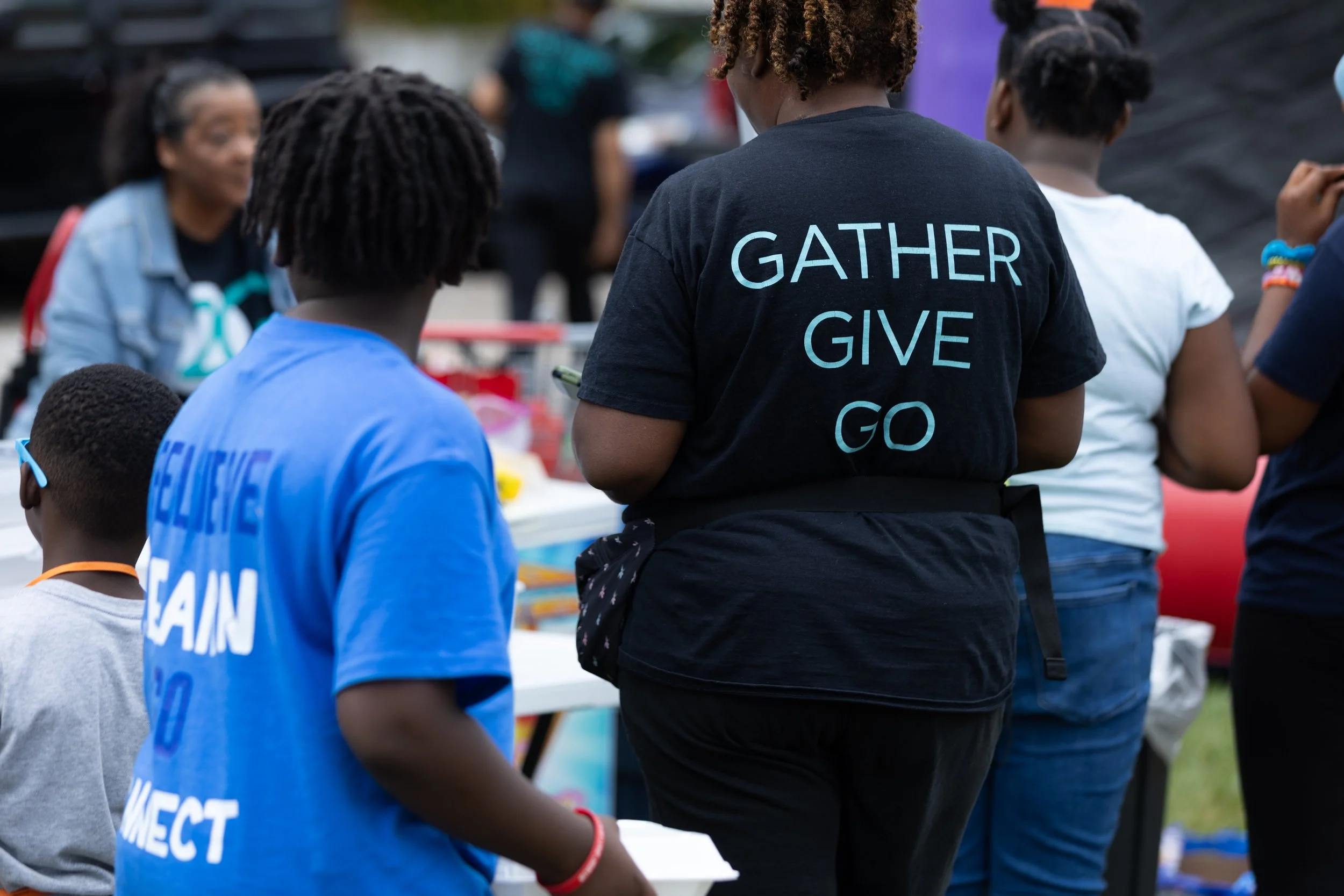 People gathered outdoors at an event or market, with one person wearing a black T-shirt that says 'Gather Give Go' on the back, surrounded by children and other attendees.
