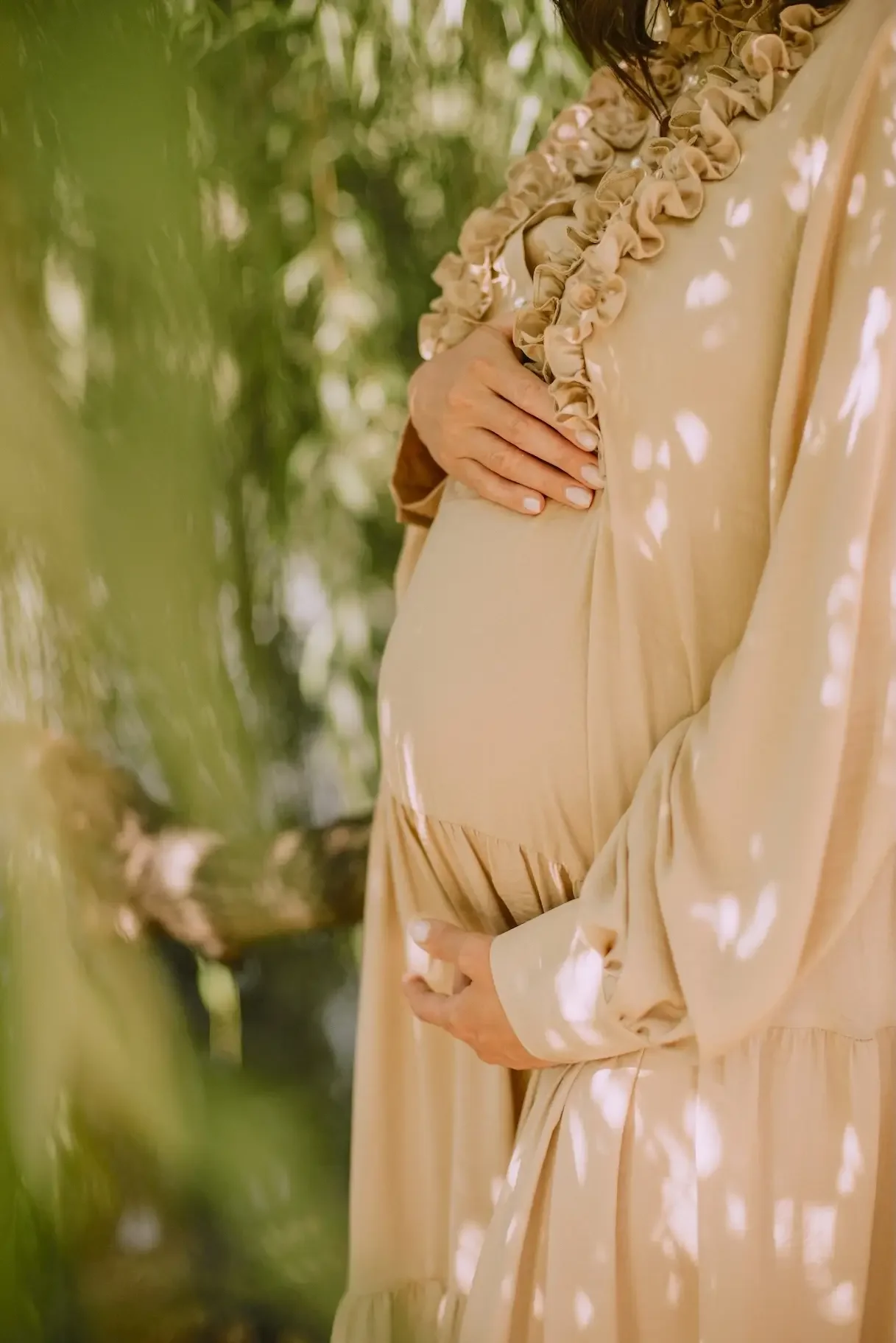 A pregnant woman in a beige dress with ruffled collar, standing outdoors among lush green trees, gently resting her hand on her belly.