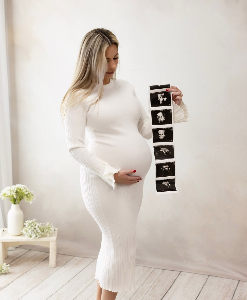 Pregnant woman in white outfit holding ultrasound images inside a room with light-colored walls, a small table with white flowers, and a wooden floor.