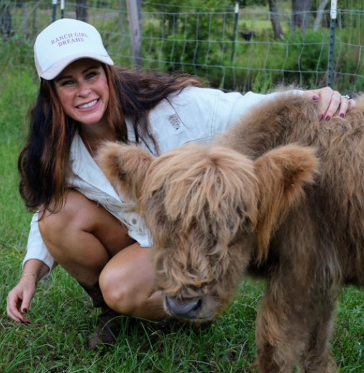 A woman crouching on the grass petting a small brown cow with a shaggy coat, in a fenced outdoor area with trees.