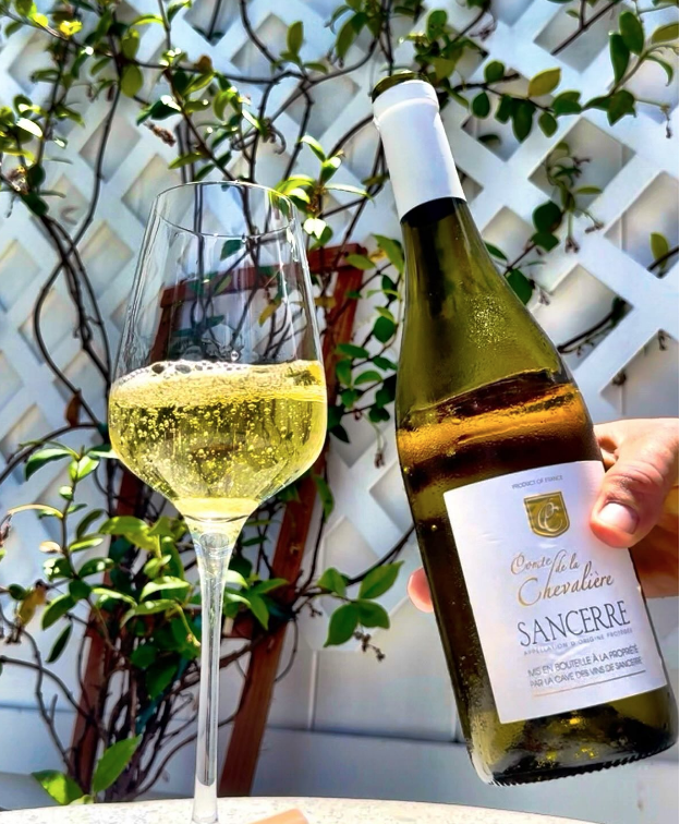 A hand holding a bottle of Samberre Sancerre wine next to a filled wine glass outdoors with green leafy plants and a white trellis in the background.
