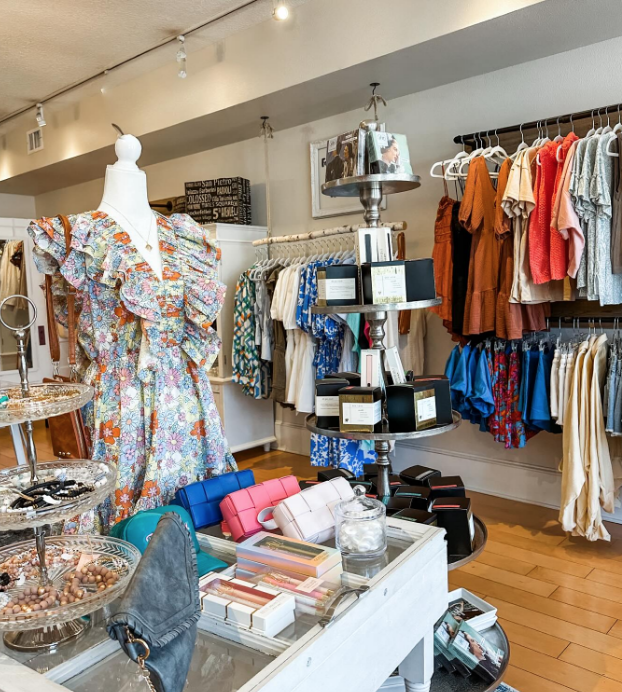Clothing boutique display with mannequins wearing floral dresses, racks of clothes in various colors, and accessories like handbags and jewelry.